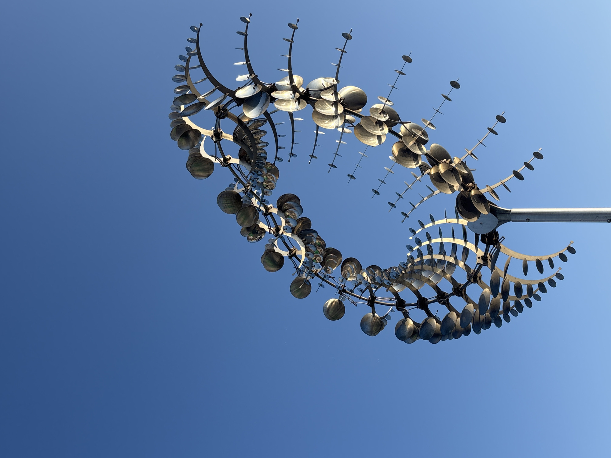 A large, kinetic metal sculpture is captured against a clear, bright blue sky in Chaikovsky, Russia. The sculpture consists of numerous reflective metallic components arranged in a complex, undulating, spiral or wave-like form, supported by a thick, light-colored vertical pole extending from the bottom of the frame. The components include larger, partially spherical or cupped elements, and smaller, flat, disc-like elements attached to thin rods. Sunlight reflects off the metallic surfaces, creating varying highlights and shadows across the structure. No people, other objects, or environmental details are present beyond the sculpture and the cloudless sky.