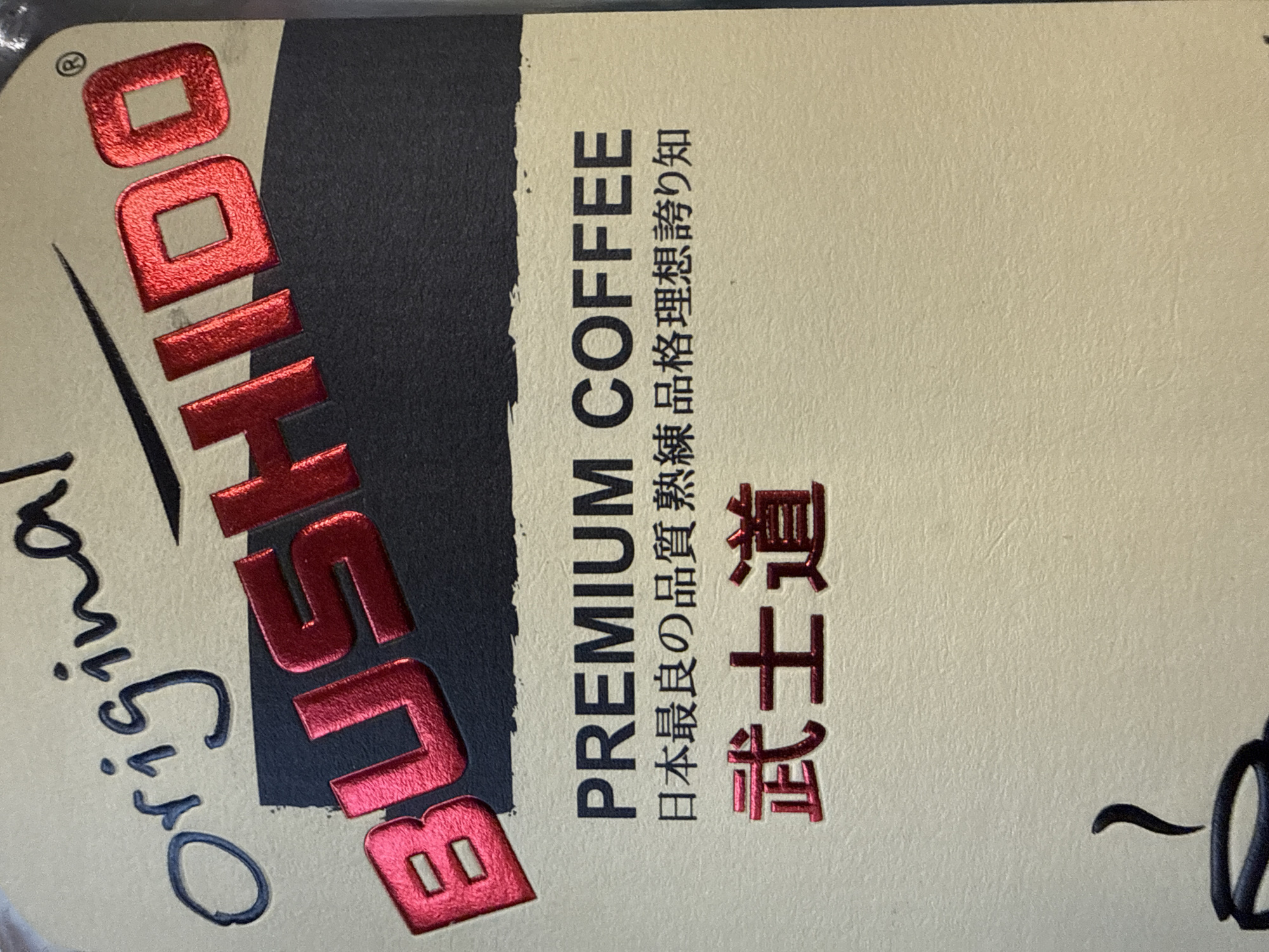 The image is a close-up of a coffee product label. The text "Original BUSHIDO" is prominently displayed in a stylized font. Below this, "PREMIUM COFFEE" is written in block letters, followed by Japanese characters. The word "武士道" (Bushido) is also displayed in large, red, metallic letters. The background is a textured, light beige color with a darker rectangular area behind the "BUSHIDO" text. There are no people or environmental elements visible that would indicate a location in Norilsk, Russia.