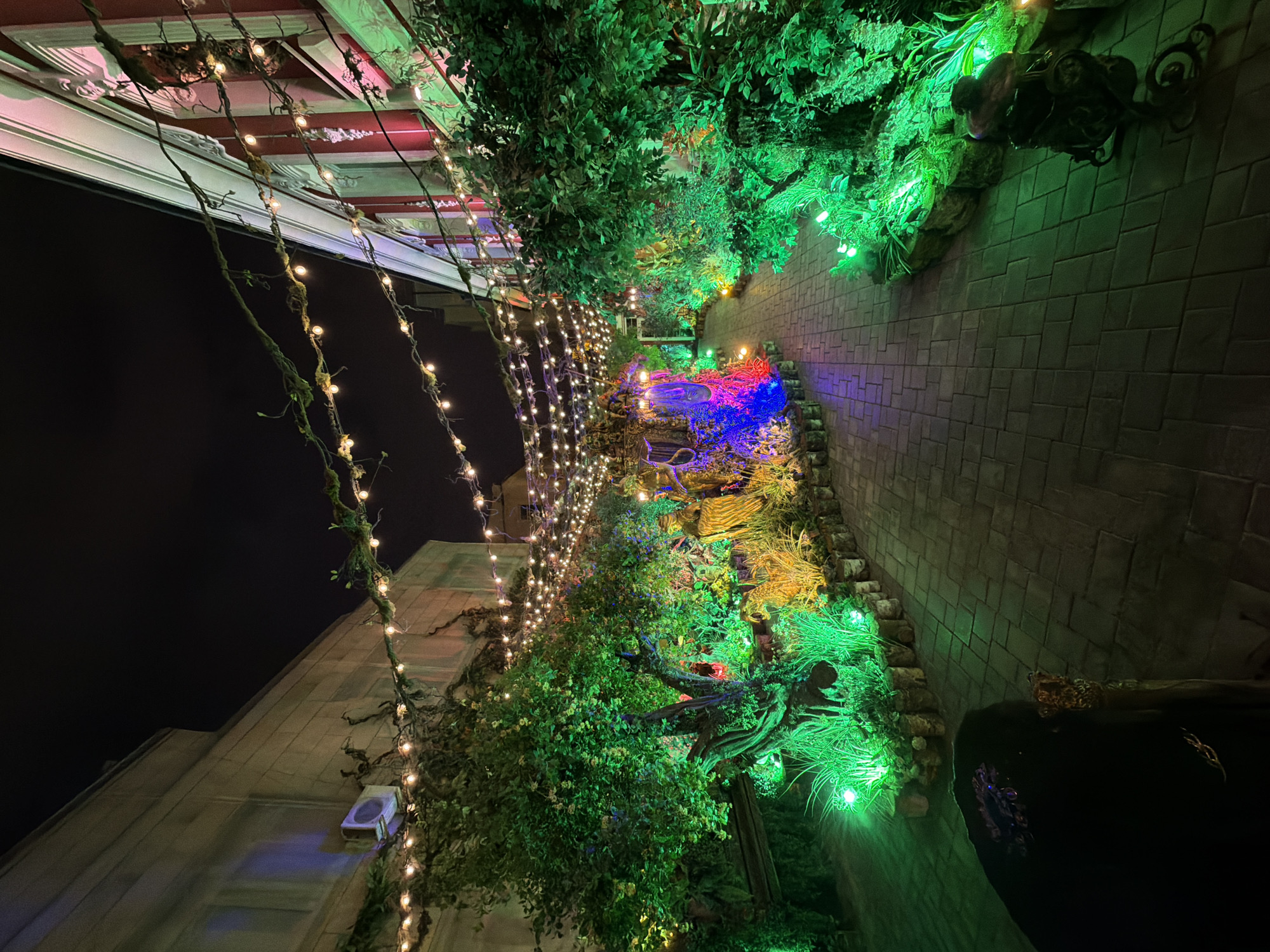 At night, a building in Moscow, Russia, is decorated with lights and greenery. The facade of the building, visible on the left side of the image, is adorned with strings of small, warm-toned lights. The structure has a light-colored exterior and an air conditioning unit is visible. Above the building, there is a roof with decorative moldings and vines.
To the right of the building, there is a vertical garden or wall with dense foliage that is illuminated with green lights. Within this garden, there is a water feature that is illuminated with blue and purple lights. The wall is constructed of brick or stone.
A person wearing dark clothing is visible in the bottom right corner of the image. The scene suggests a festive or celebratory atmosphere.