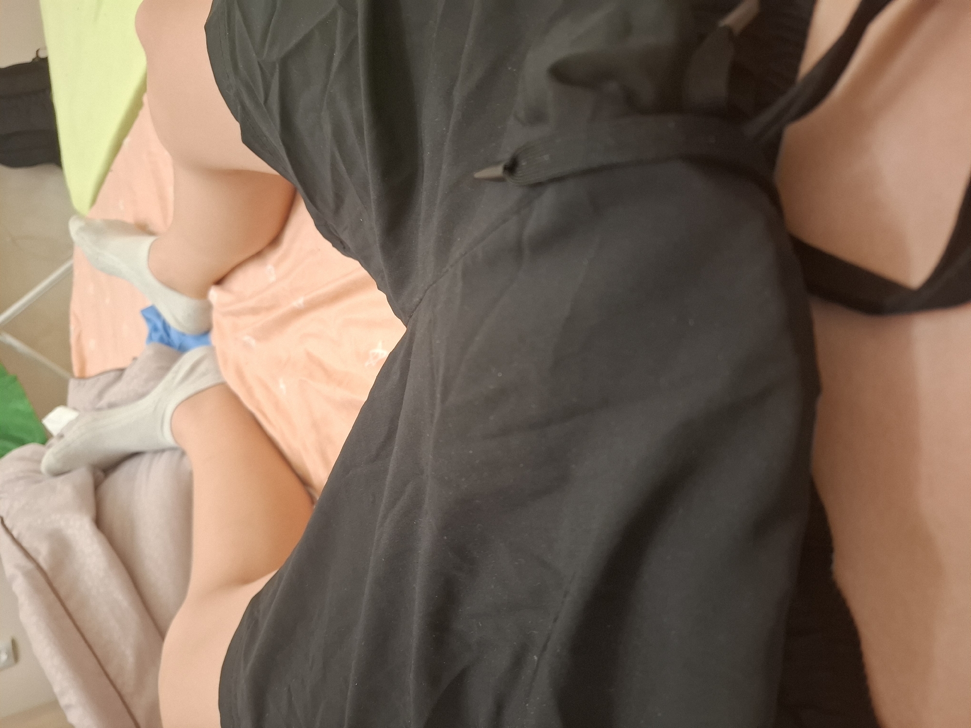 A close-up, high-angle shot depicts a person lying on a bed. They are wearing gray socks and a black garment, possibly a dress or a robe. The bed is covered with light pink and light green sheets, and a gray blanket is visible on the left side. The scene does not provide enough information to determine the location in Yambol, Bulgaria.