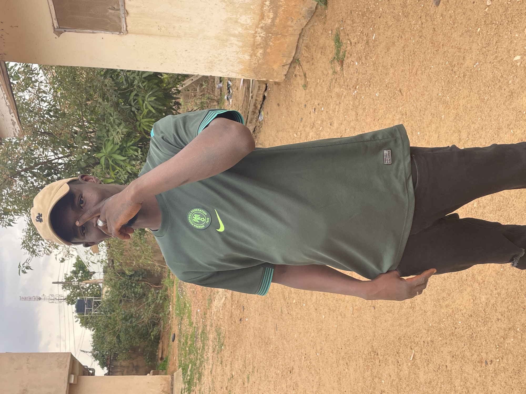 A young man stands on a dirt surface in Jos, Nigeria. He is wearing a khaki cap, a green short-sleeved t-shirt with a Nigeria Football Federation logo and teal trim, and dark pants. He is holding one hand up to his face. Buildings and trees are visible in the background, with a communications tower rising above the trees.