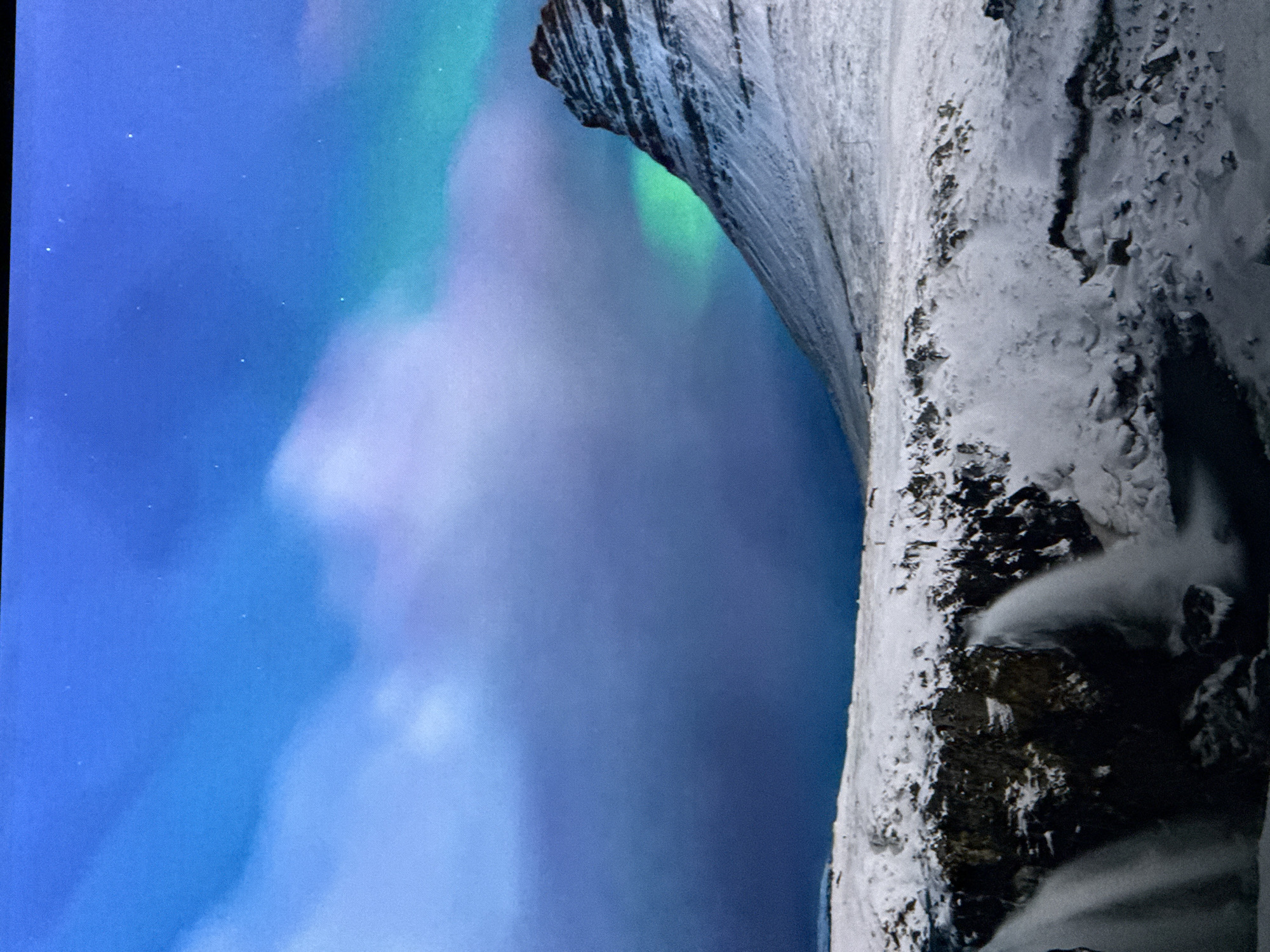 The image depicts a dramatic natural scene, likely a nighttime landscape. The left side of the frame is dominated by a deep blue sky, speckled with faint stars. A vibrant aurora borealis, or Northern Lights, stretches across the sky in swirling bands of green and purple. The aurora illuminates the sky with ethereal light, creating a magical atmosphere.
On the right side of the image, a steep, snow-covered rock formation rises vertically. The rock face is textured and rugged, with dark crevices and patches of snow clinging to its surface. The snow appears to be freshly fallen and thick, suggesting a cold, wintry environment. The contrast between the dark, solid rock and the soft, luminous aurora is striking.
The orientation of the image is vertical, with the sky and aurora appearing on the left and the rock formation on the right, creating a sense of immense scale. The lighting suggests it is night, and the presence of the aurora indicates a location in a polar region. The overall mood is one of awe and wonder, capturing the beauty and power of nature. There is no visible text in the image.