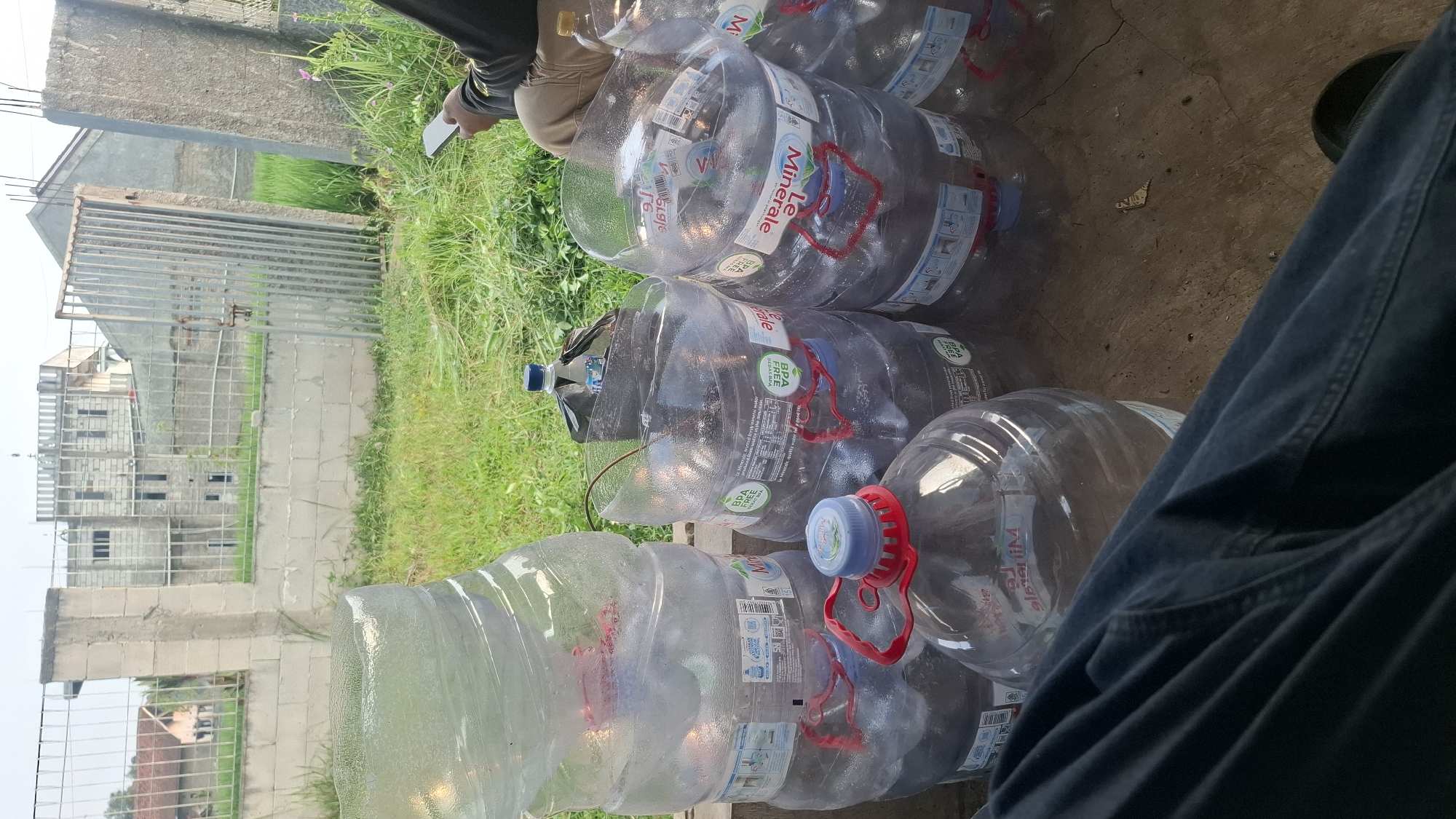 In Rahong, Indonesia, a scene unfolds with several large, clear plastic gallon water jugs, some full and some partially empty, arranged on a cracked concrete surface. The jugs are branded with "Le Minerale" and "Le Minerale BPA FREE," indicating they contain mineral water. Some of the jugs have red handles attached to their necks. In the background, a metal gate with vertical bars stands partially open, leading to a grassy area. Beyond the gate, a building under construction with visible rebar and concrete walls is present, along with a smaller, older-looking house. A person, whose legs and hip are visible in the foreground wearing dark blue denim jeans, is seated, holding a white smartphone in their right hand, suggesting they are interacting with it. The overall environment appears to be an outdoor, possibly semi-abandoned or construction site area, with overgrown grass and scattered debris.
