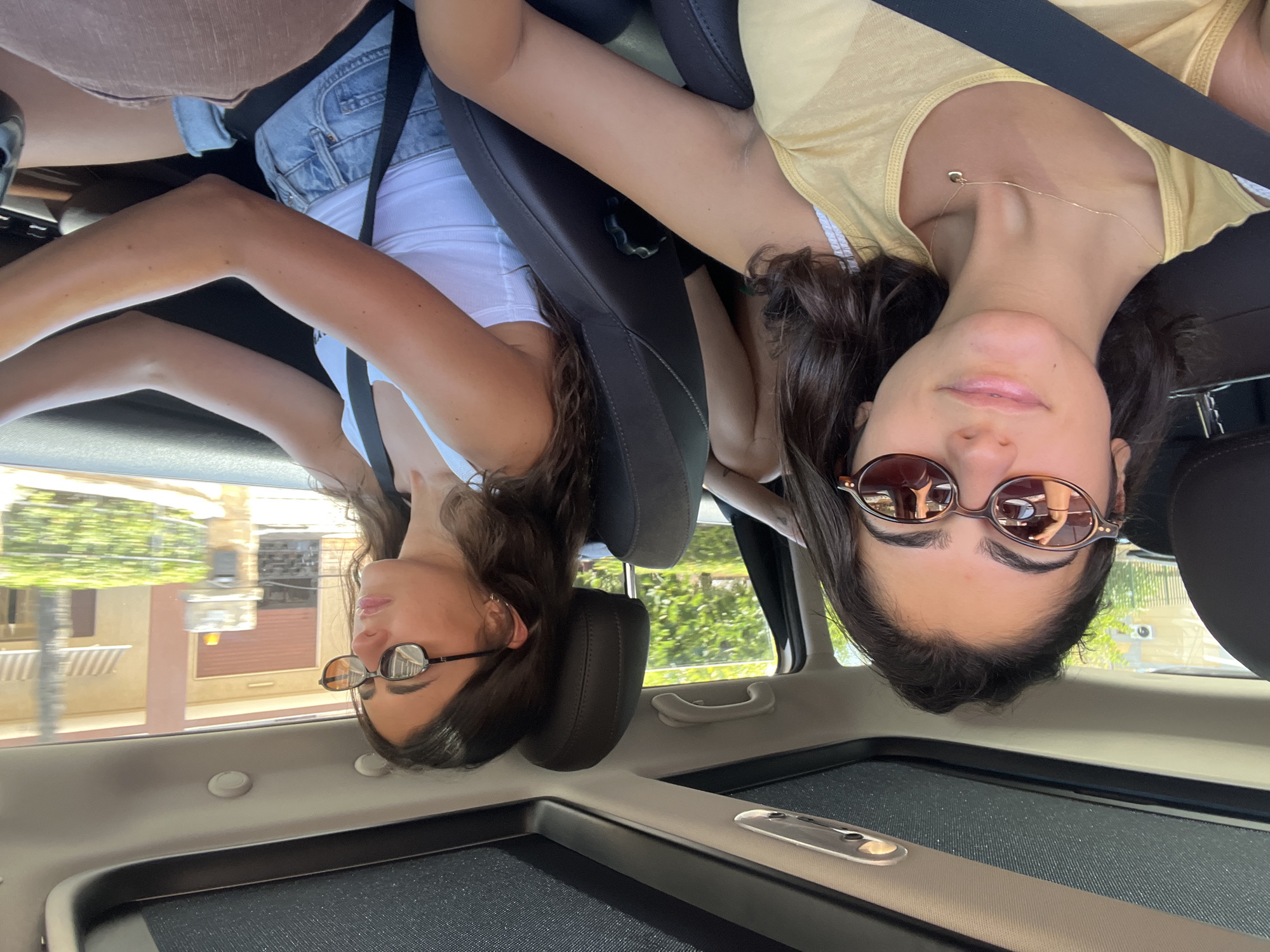 Two young women are inside a car, with one in the driver's seat and the other in the passenger seat. The woman in the passenger seat is closer to the camera and looking at it, while the woman in the driver's seat is looking forward, presumably at the road. Both are wearing sunglasses and casual clothing. The car's sunroof is open, revealing a bright sky. Outside the car, greenery and buildings are visible, suggesting an urban or suburban environment. The image appears to be taken in Fasano, Italy, as indicated by the style of the buildings and vegetation.