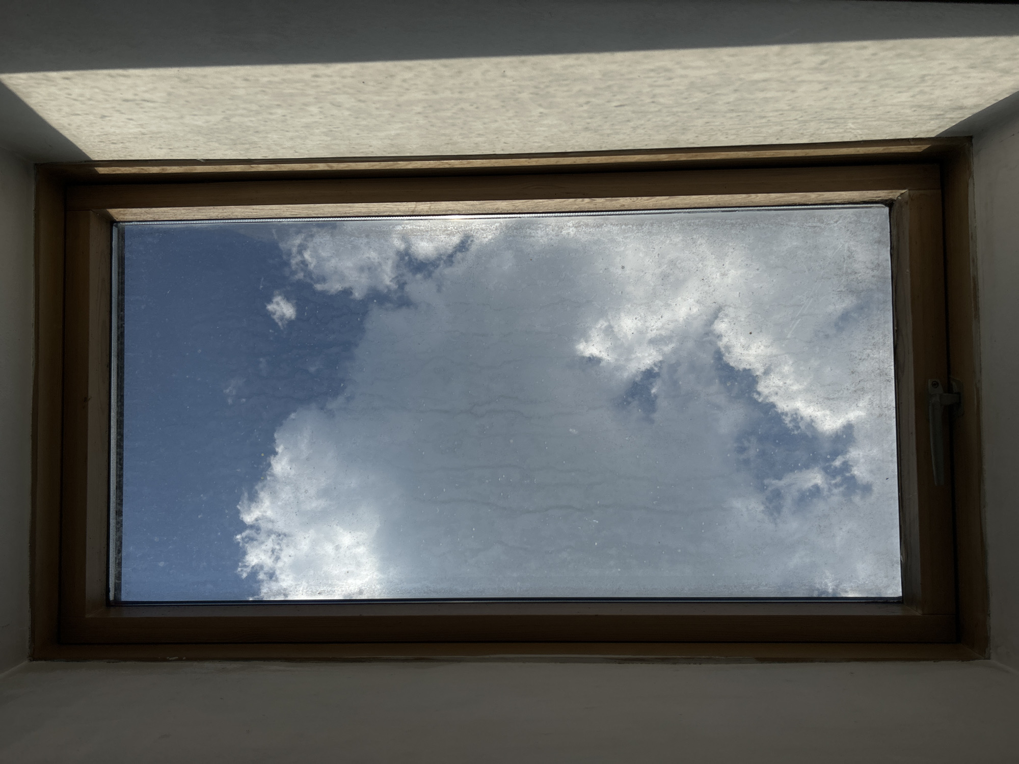 The image captures a view of a blue sky with fluffy white clouds through a window. The window has a wooden frame and appears to be a skylight or an overhead window, given the angle of the light casting a shadow on the ceiling-like structure above. The glass of the window shows some streaks or smudges, particularly in the upper half. Sunlight is filtering through the clouds, creating bright patches and casting shadows, suggesting it is daytime. The overall scene is serene and peaceful, evoking a sense of looking up at the sky. There are no people or specific objects other than the window and the sky. The setting appears to be indoors, looking outwards. There is no visible text in the image.