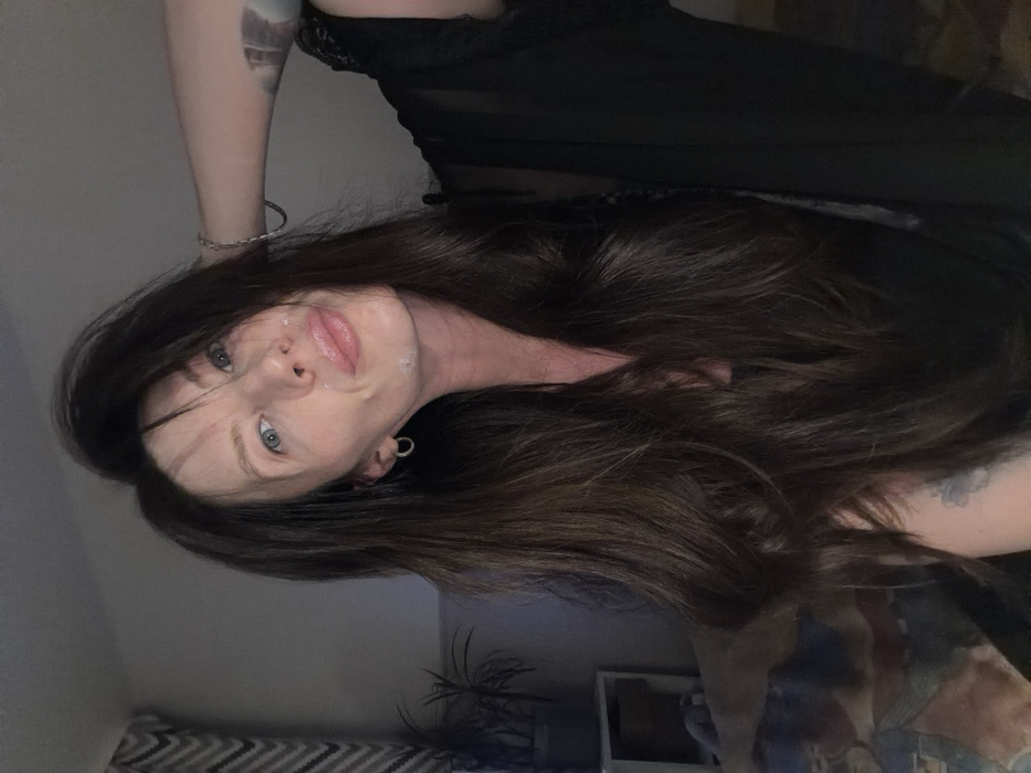 A person with fair skin and long, dark hair looks towards the camera with a slight smile. The individual has blue eyes and wears a dark, possibly lace-trimmed garment. A circular earring is visible on the left ear. Light appears on the right side of the face, contrasting with darker tones on the left. A portion of an arm with a tattoo is visible. A plant and storage unit are shown in the background.