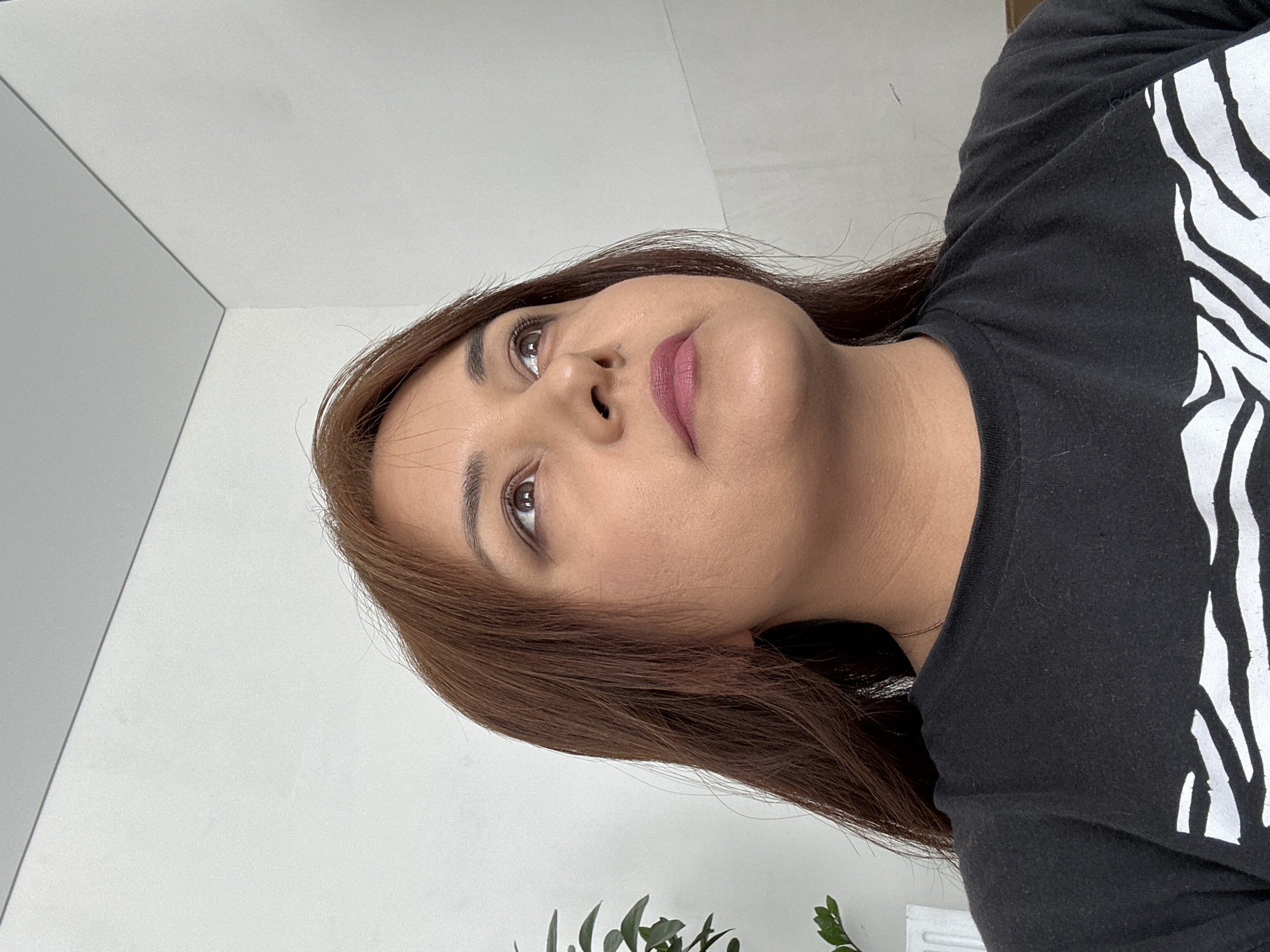 A medium shot depicts a woman looking upwards. She has light brown hair and fair skin, with noticeable makeup on her lips and eyes. She is wearing a dark t-shirt with a white pattern on the upper right side. The background includes a white wall on the left, and what appears to be a plant in the lower center. The location is Unknown, Unknown.