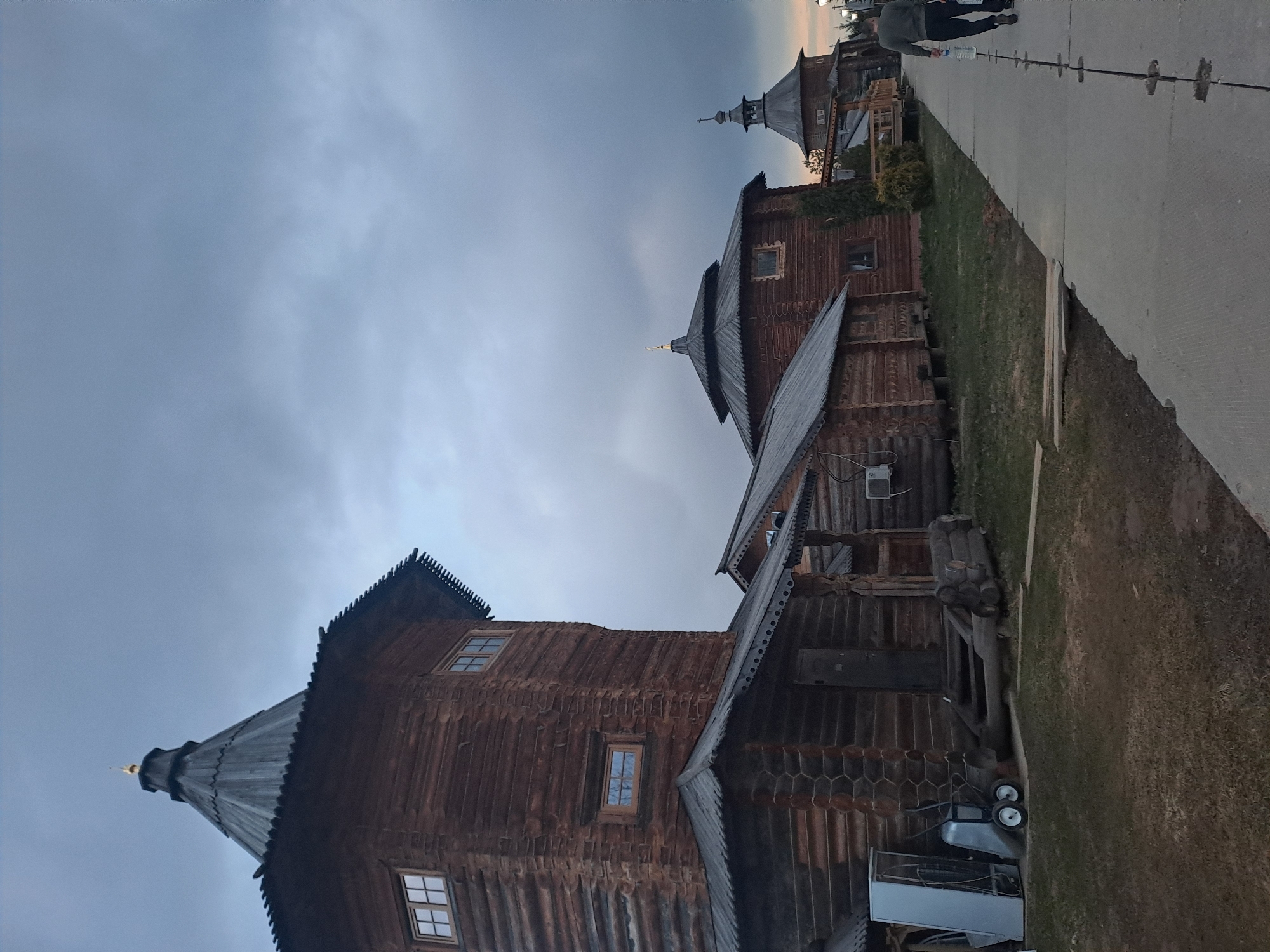 The image shows wooden buildings in Vzglyadnevo, Russia. The buildings are constructed with dark wood, featuring pointed roofs and small windows. The sky is cloudy and grey. A paved pathway is present with grass alongside it, and individuals are walking on the pathway.
