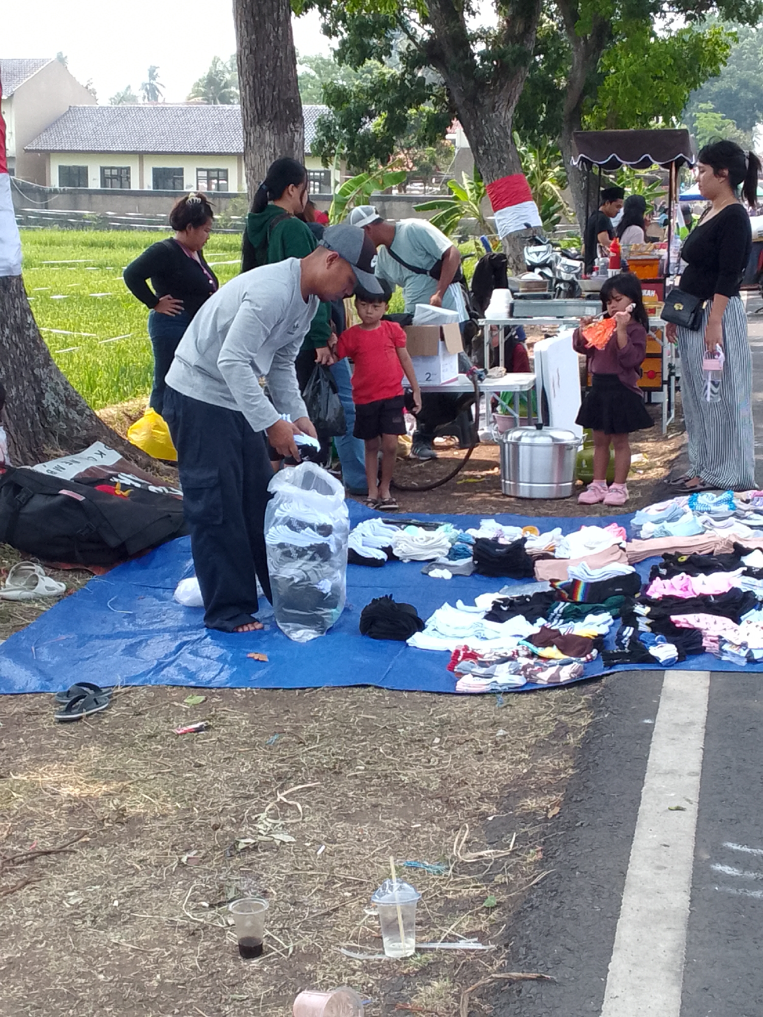 This image captures an outdoor market scene in Mandalawangi, Indonesia. A man in a grey long-sleeved shirt and dark pants is seen in the foreground, sorting through clothes placed in a large clear plastic bag. He is standing on a blue tarp spread on the ground, which also displays a variety of folded clothing items, socks, and shoes. To the left of the man, a large black duffel bag and a pair of white sandals are visible. In the background, several people are gathered around stalls set up under trees. A food stall with various food items and a steam cooker is also present. The environment appears to be a street or roadside, with a grassy area on one side and the paved road with a white line on the other. The overall atmosphere suggests a casual local gathering or bazaar.
