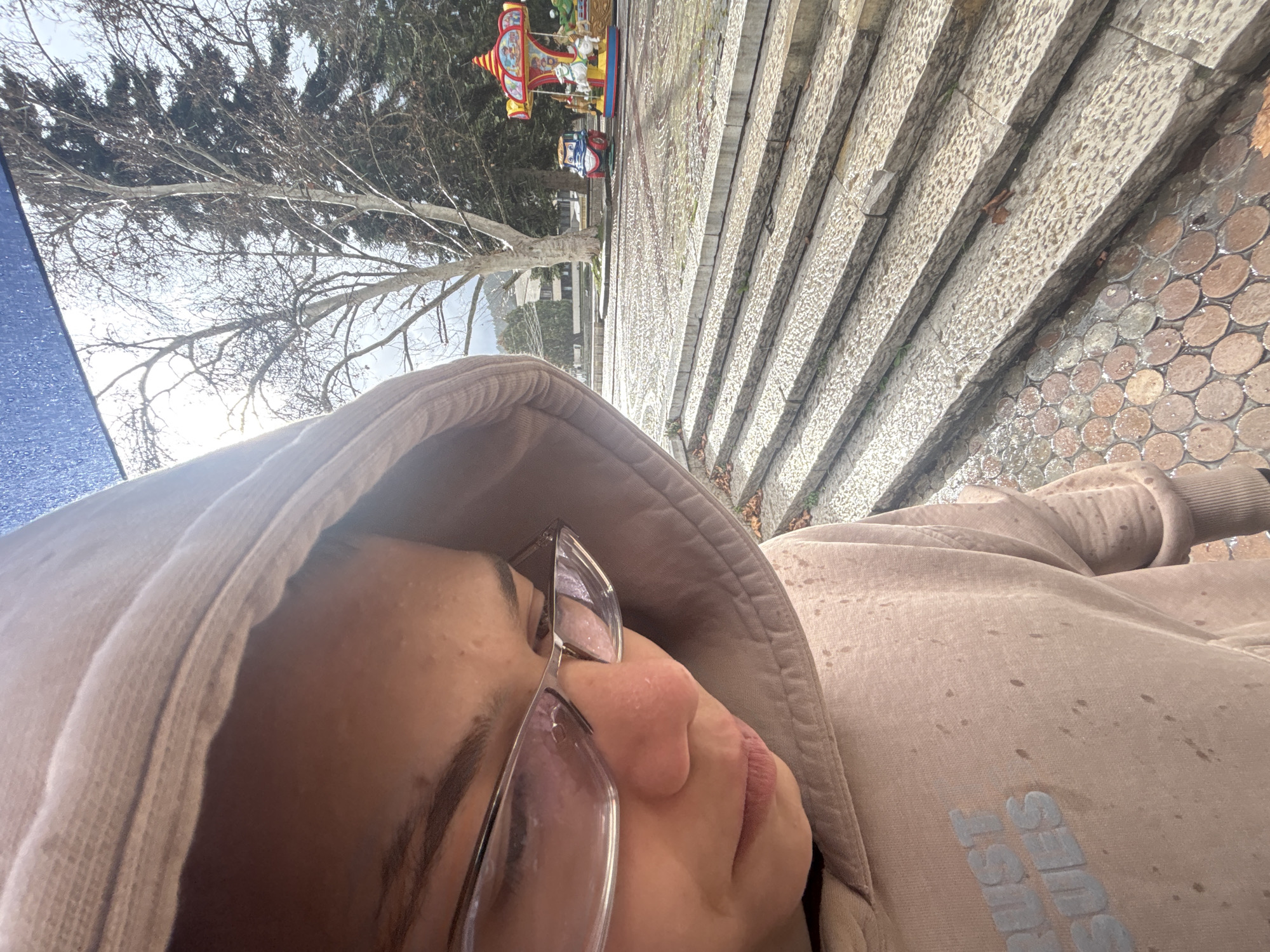 An individual is captured in a close-up, downward-angled shot, partially obscuring the wider scene. The person, viewed from the upper torso and head, wears a light brown or beige hooded sweatshirt with the hood up, covering most of the hair. Clear-framed glasses rest on the bridge of the nose, and the gaze is directed downwards and slightly to the right. The visible portion of the sweatshirt's back displays light-colored text that appears to read "RUST ISSUES" and is speckled with numerous small, dark brown droplets or stains. A blue object, possibly an umbrella, is visible in the upper left of the frame, partially reflecting on the person's hood.
The background reveals an outdoor urban environment, specifically a paved public square or park in Troyan, Bulgaria. The immediate midground features a series of wide, shallow, light grey stone steps leading down to a wet, paved area. This area is patterned with light-grey textured paving stones, and to the lower left of the steps, a section of reddish-brown circular pavers is visible.
Further into the background, two children's coin-operated rides are present: a red and blue train-like vehicle with a smiling face, and a brightly colored carousel featuring white horses. Behind these rides stands a large, bare deciduous tree with light bark, indicative of late autumn or winter, and to its right, a dark green evergreen tree. In the far background, a multi-story building with light facades and dark windows is discernible, with distant mountains or hills rising behind it. The sky is bright and overcast, and the wet ground suggests recent precipitation.
