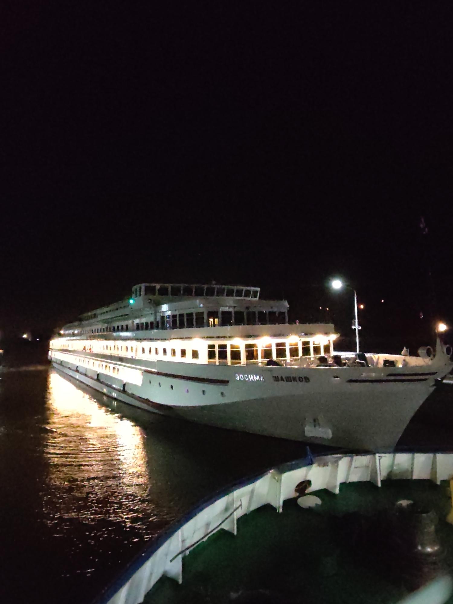 At night in Iksha, Russia, a multi-deck river cruise ship, "Zosima Shashkov," is docked next to another ship. Lights illuminate the "Zosima Shashkov," reflecting on the water. The focus is on the moored ship, while the surrounding environment is dark.