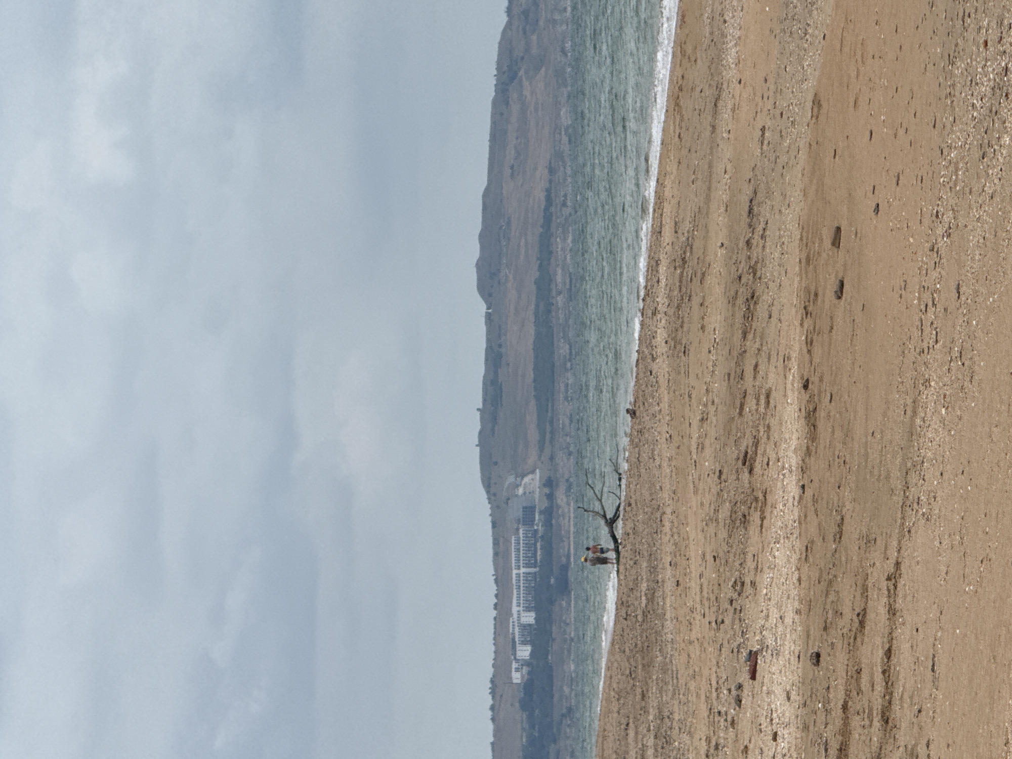 A distant view of a beach scene in Kerch, Ukraine, shows two individuals standing on the shore near a piece of driftwood. The beach is composed of sand and small stones, with the sea on the right side of the image. In the background, a large, white building complex is visible on a hill, with a smaller structure on a higher peak. The sky is overcast with gray clouds.