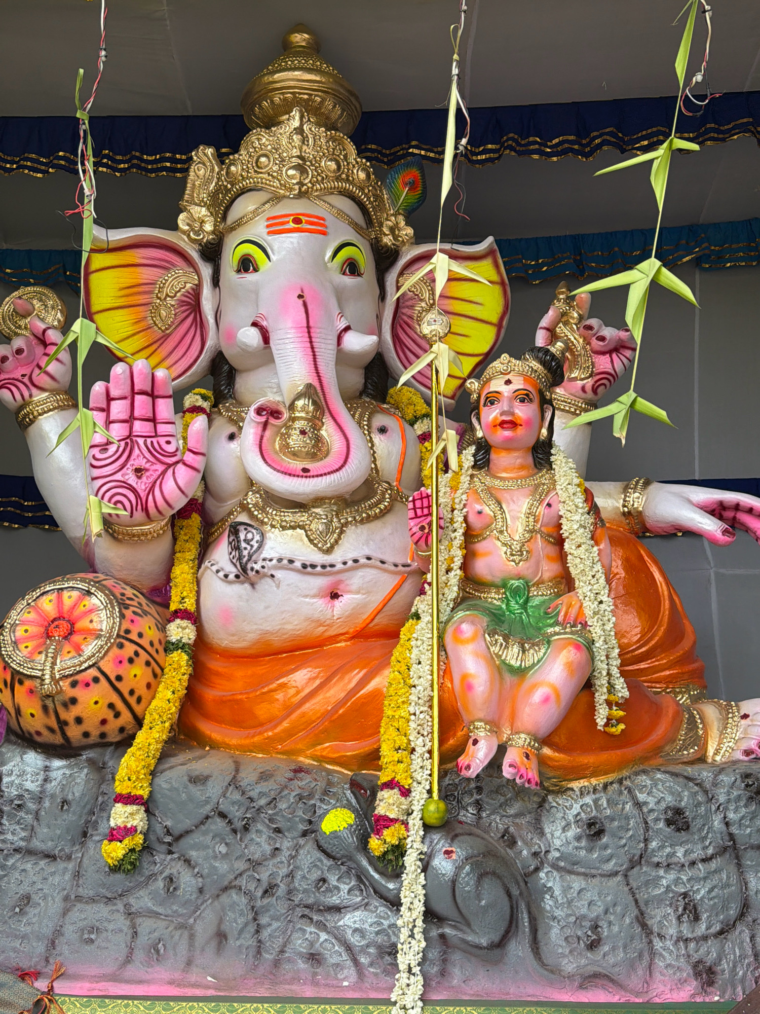 The image depicts a religious idol of Lord Ganesha and a child deity, likely Murugan, seated on a grey, rocky platform. The idols are adorned with garlands of yellow and white flowers. The larger idol of Ganesha is white with pink accents and wears an elaborate golden crown. The child deity is pink and green, also wearing a golden crown and intricate jewelry. In the background, a blue and gold fabric drapes across a tent-like structure. Green palm fronds are suspended from above by strings. A decorated orb sits to the left of Ganesha. The scene appears to be an outdoor religious display, possibly during a festival, in Pallavaram, India.