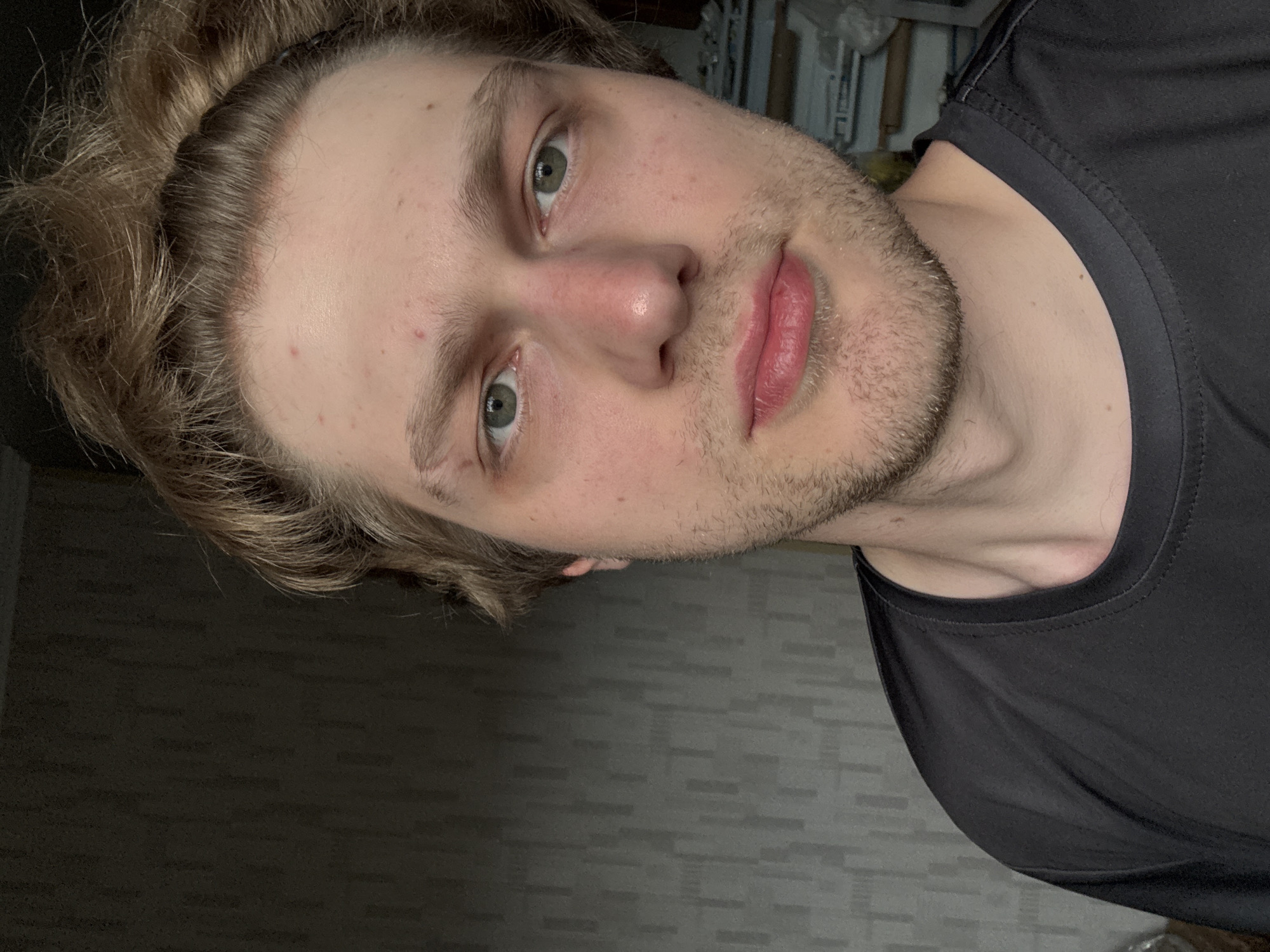 The image is a close-up selfie of a young Caucasian man, taken indoors. He has light brown hair styled upwards, green eyes, and fair skin with some redness on his cheeks and nose. He has stubble on his chin and upper lip. He is wearing a black t-shirt. The background is blurred, with a textured wall on the left and some indistinct objects on the right, possibly shelves or storage. The lighting appears to be natural, coming from the left. There is no discernible location in the city of Khimki, Russia, or any specific context provided by the image itself.