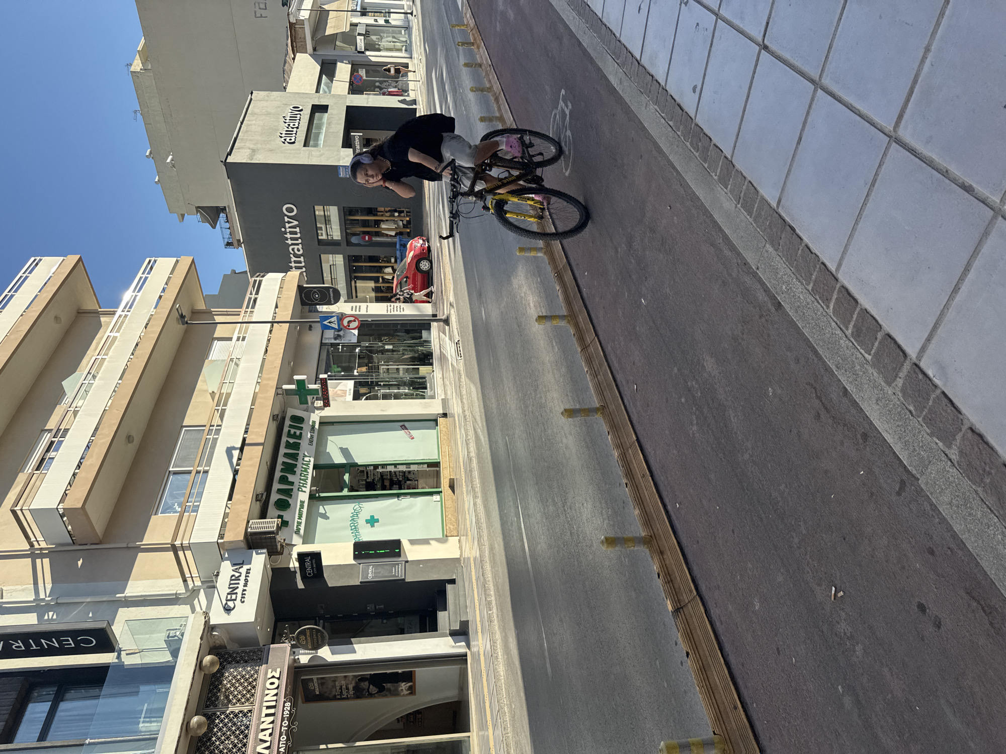 The image captures a street scene in Chania, Greece, likely on a sunny day. In the foreground, a person is riding a bicycle on a dedicated bike lane that runs alongside the road. The cyclist, wearing a black t-shirt and light-colored shorts, has headphones on and appears to be touching their ear. The bicycle is dark-colored with yellow accents and pink shoes are visible on the rider's feet.
The street itself is paved, with a red-colored bike lane separated from the main road by yellow bollards and a wooden barrier. On the right side of the road, a sidewalk with large square tiles is visible.
The background features buildings lining the street. On the left, a building with the sign "CENTRAL CITY HOTEL" is prominent, with shops below. To the right, other commercial establishments are visible, including one with a green facade and the word "PHARMACY" in English and Greek. Further down the street, more buildings and shops are present, with one displaying the sign "attrattivo". A red car is parked on the street. The sky is clear and blue. The overall scene depicts a typical urban environment in Chania.