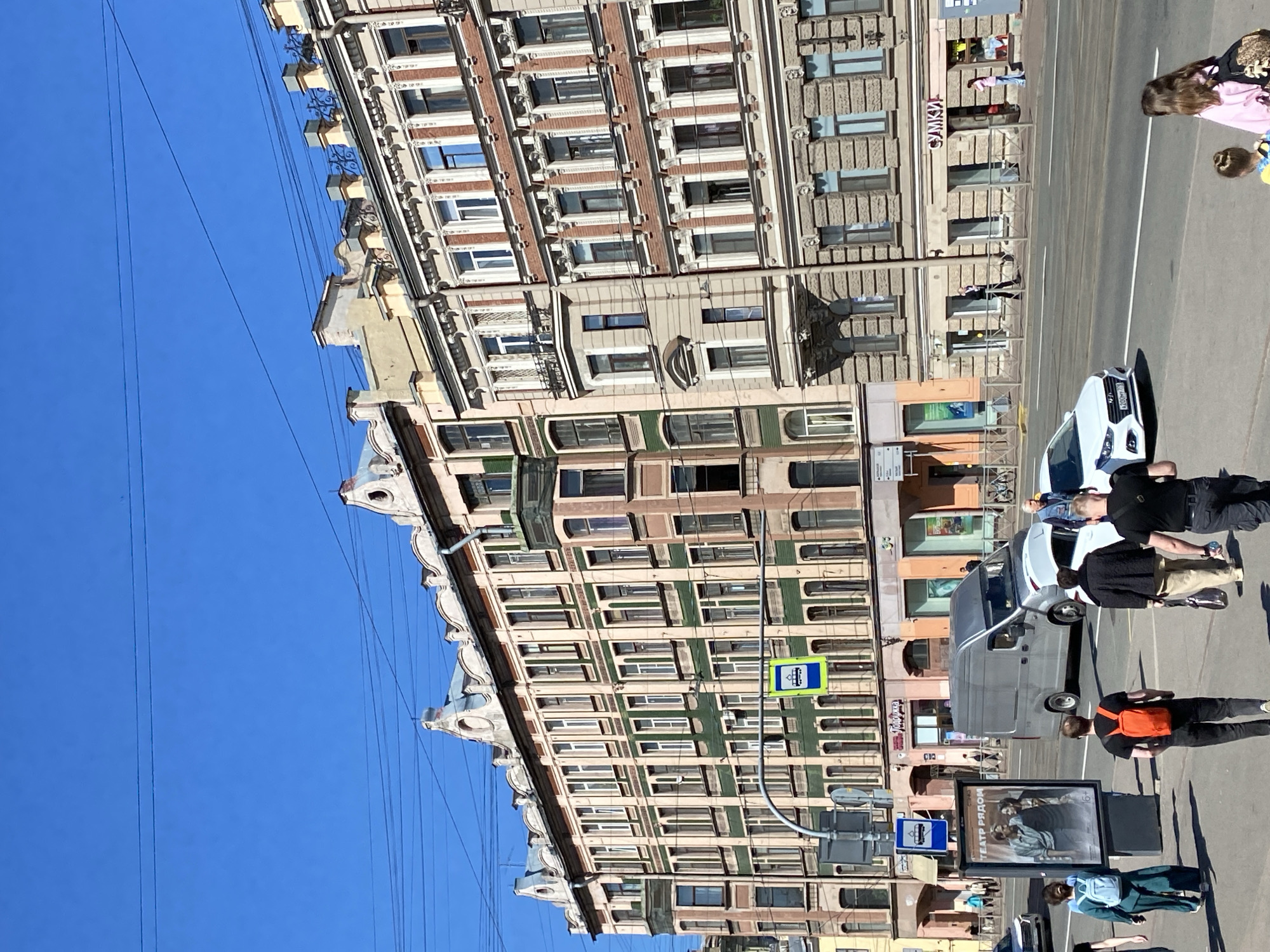 The image presents a sunlit street scene in Saint Petersburg, Russia, showcasing ornate historical architecture, pedestrians, and vehicles under a clear blue sky. The original image is rotated, but the description assumes an upright perspective where buildings are vertical and the street is at the bottom.
Two prominent multi-story buildings line the street. The one on the left features a distinctive facade with alternating sections of light beige and dark green, punctured by numerous windows. Its upper levels are adorned with elaborate cornices and decorative brackets. Stretching across the bright blue sky above are many thin overhead wires, indicative of public transport like trams or trolleybuses common in the city.
Further down the street, another grand building displays a facade of reddish-brown brickwork interspersed with grey sections. This structure also boasts numerous windows and decorative architectural details. A shop sign on its ground floor reads "САНЫЧ" (SANЫCH).
The street itself is wide, with white lane markings visible on the asphalt. Several vehicles are present, including a white sedan (its license plate partially visible as "X208MM") and a dark grey van. Pedestrians are scattered across the scene; some are walking on the pavement, while others are near the vehicles. In the foreground, a person wearing an orange backpack walks past an A-frame advertising sign. This sign features an image of several people and bears the Russian text "ТЕАТР РЯДОМ" (TEATR RYADOM), which translates to "Theater Nearby".
Blue and yellow bus stop signs, depicting a bus icon, are visible on poles along the street. The bright sunlight indicates a clear and pleasant day, likely midday or early afternoon, with distinct shadows cast by the buildings and street furniture. The scene captures a typical bustling street environment in the historic city.