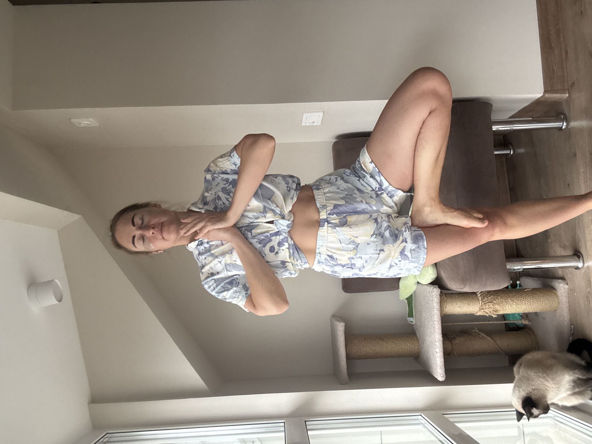 A woman in a patterned blue and white short-sleeve top and shorts sits on a grey chair near a window, her right leg extended and resting on the chair while her left leg is bent with her foot tucked under her thigh. She is touching her neck with her right hand. A cat tree sits next to the chair and a Siamese cat is visible by the window. The room is indoors, likely in a residential area. The location, Chaikovsky, Russia, is a speculative geographic detail.