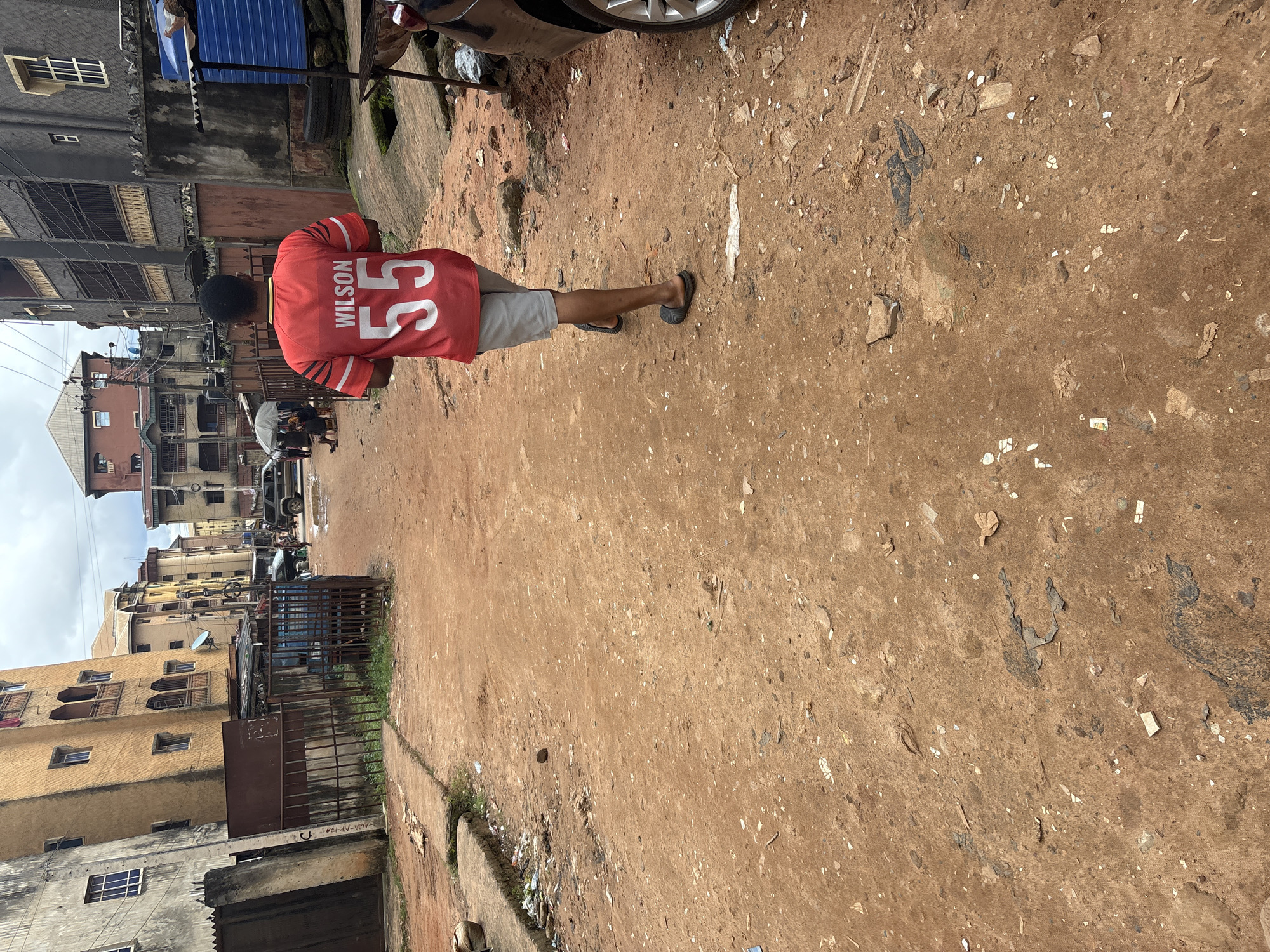 A young Black male, wearing a red t-shirt with "WILSON 55" printed on the back in white, and grey shorts, walks away from the camera down a dirt road. He wears black slides on his feet. The road is lined with multi-story buildings on both sides. The buildings appear to be residential or commercial, with some showing signs of age and wear. There are metal fences and gates along the sides of the road. Debris and trash are scattered on the ground. The sky is overcast. The scene appears to be in a densely populated urban area. This is likely a street in the city of Onitsha, Nigeria, known for its bustling markets and varied architecture.