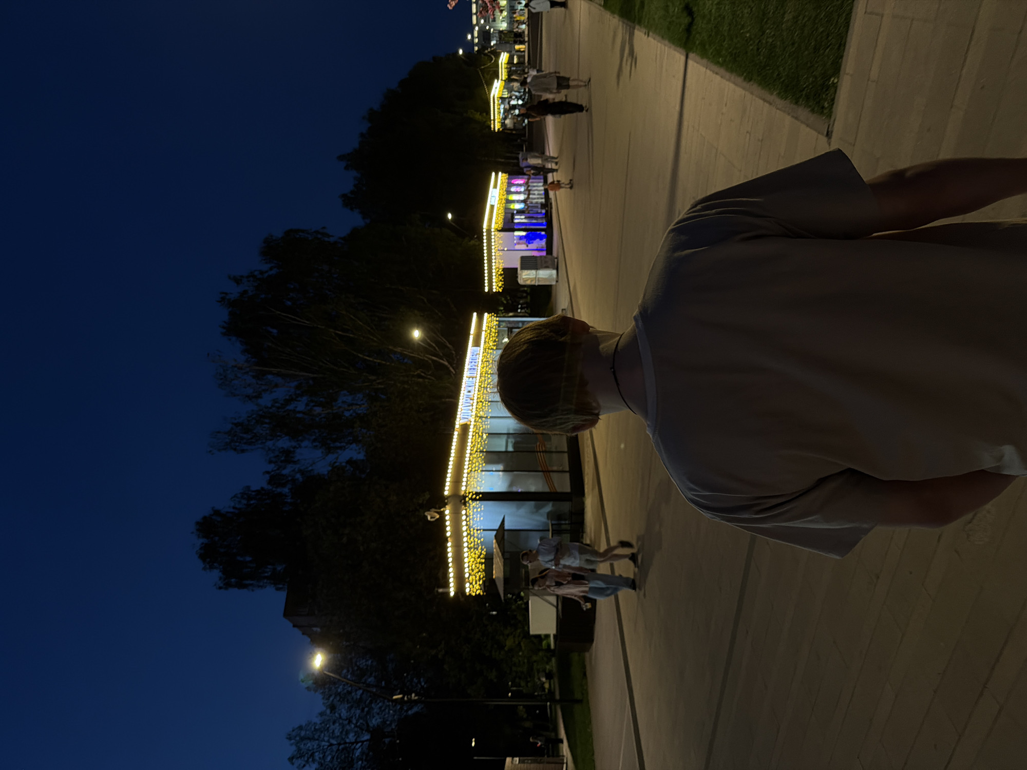 The image shows a person standing on a pedestrian walkway at night in Izhevsk, Russia. The walkway is lined with food kiosks adorned with decorative lights. Several people are walking on the walkway. The sky is dark blue above trees and buildings. A streetlamp is visible in the lower left corner.