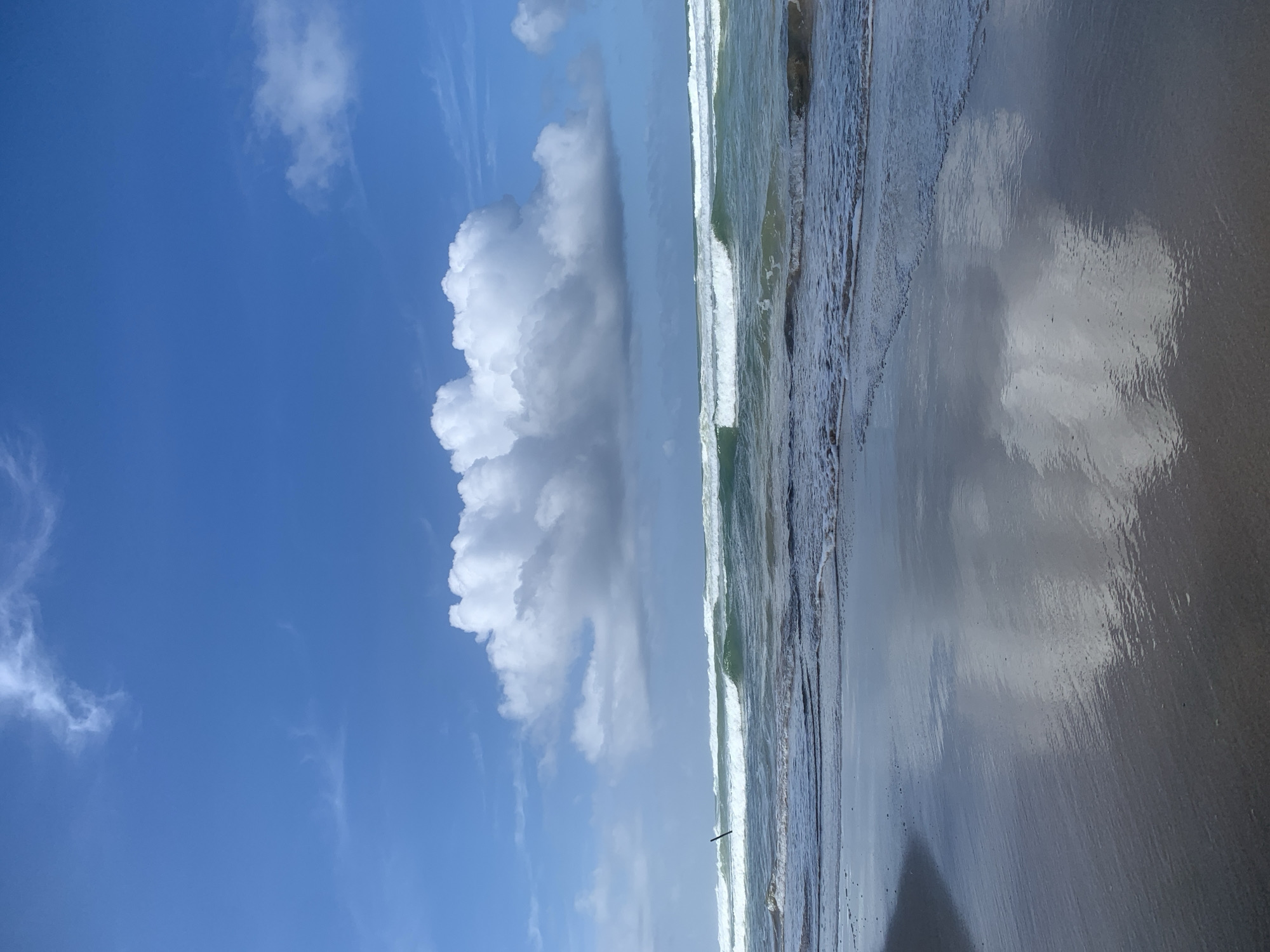 The image captures a serene beach scene in Kathaluwa, Sri Lanka. The foreground is dominated by wet sand reflecting the sky and clouds, with gentle waves lapping at the shore. In the midground, the ocean displays multiple lines of breaking waves with white foam, transitioning into a deeper blue. A thin, dark, vertical object, possibly a piece of driftwood or a marker, stands in the water on the left side. The sky is a vibrant blue, dotted with fluffy white cumulus clouds, the largest of which is positioned prominently above the horizon. The overall impression is one of a peaceful and beautiful coastal environment.