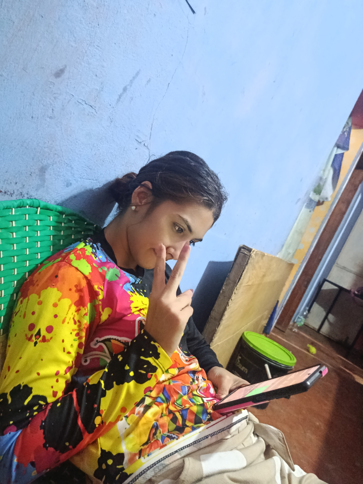 A woman is sitting indoors, possibly in a chair, holding a smartphone. She has dark hair pulled back in a bun and is wearing a colorful, long-sleeved shirt. She is looking at the phone screen. Her left hand is raised, making a peace sign. The walls are painted a light blue color with visible cracks. In the background, there is a doorway leading to another room where a box, bucket with green lid and other small objects are. There is no indication that this is in the city of Juan Griego, Venezuela.