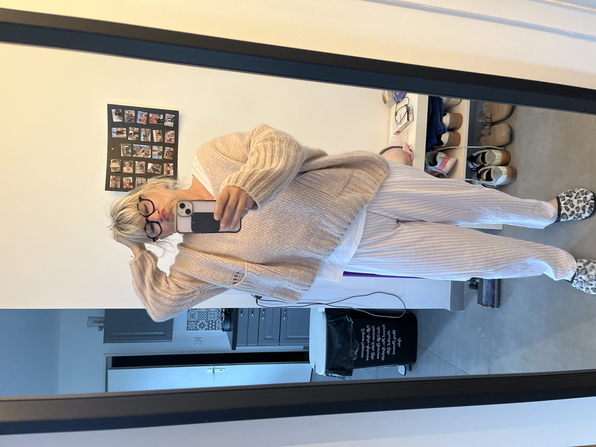A woman stands indoors taking a mirror selfie. She wears glasses, a beige sweater, and striped pajamas with leopard print slippers. In the background, a collage of photos is on the wall. A shoe rack with various shoes is visible to the right. The woman's reflection includes a glimpse of a kitchen area with cabinets and a black bag with white lettering. Based on location data from Google Maps, this image likely originates from a residence in Chambœuf, France.