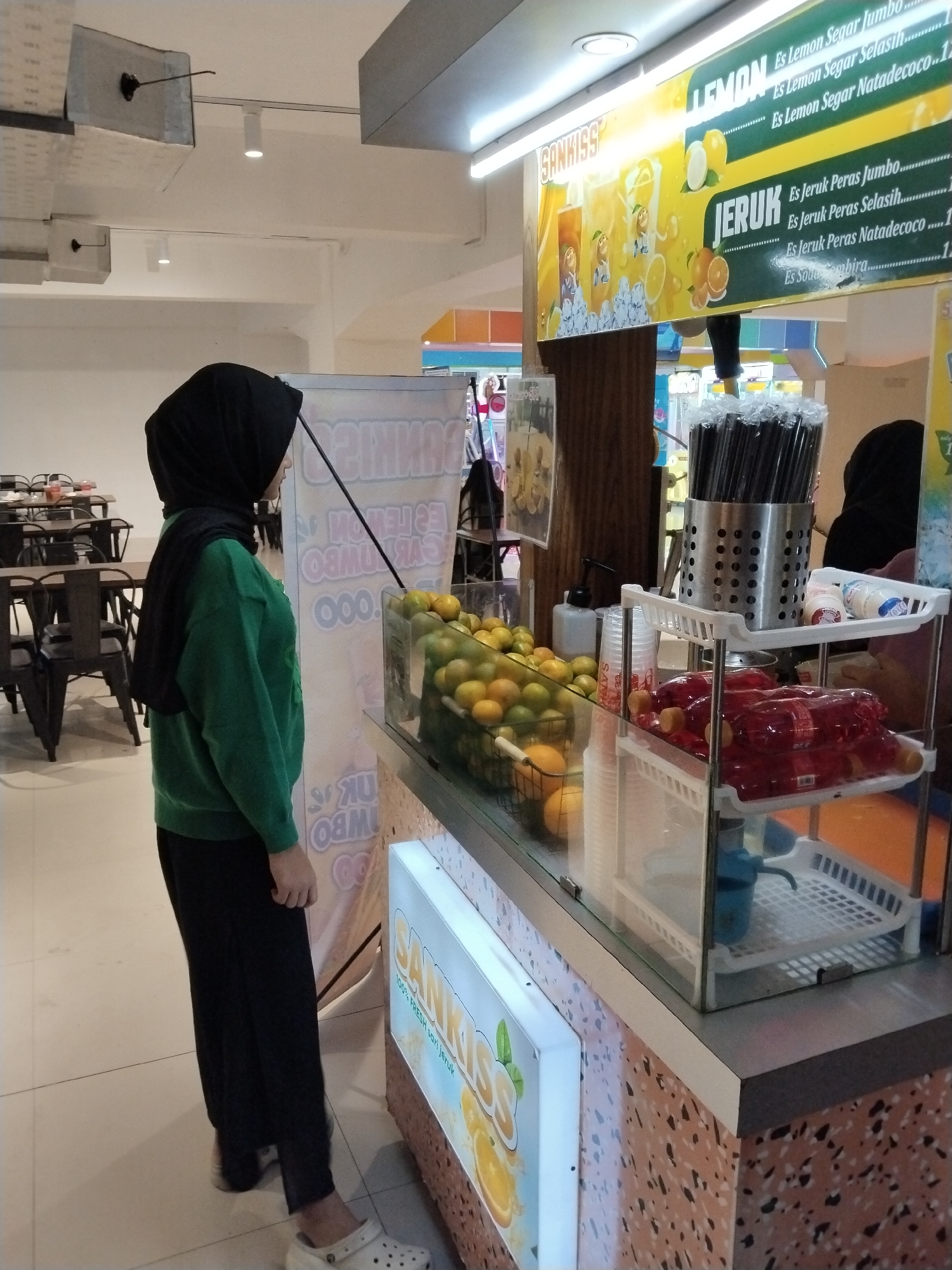 In Kecamatan Pati, Indonesia, a young woman wearing a black hijab and a green long-sleeved shirt stands beside a juice stand. The stand displays various fruits, including oranges, and bottled drinks. A sign above the stand advertises "SUNKISS" beverages, with text in Indonesian indicating flavors like "Es Lemon Segar" (Fresh Lemon Juice) and "Es Jeruk Peras Jumbo" (Jumbo Squeezed Orange Juice). The environment appears to be an indoor market or food court, with tables and chairs visible in the background.