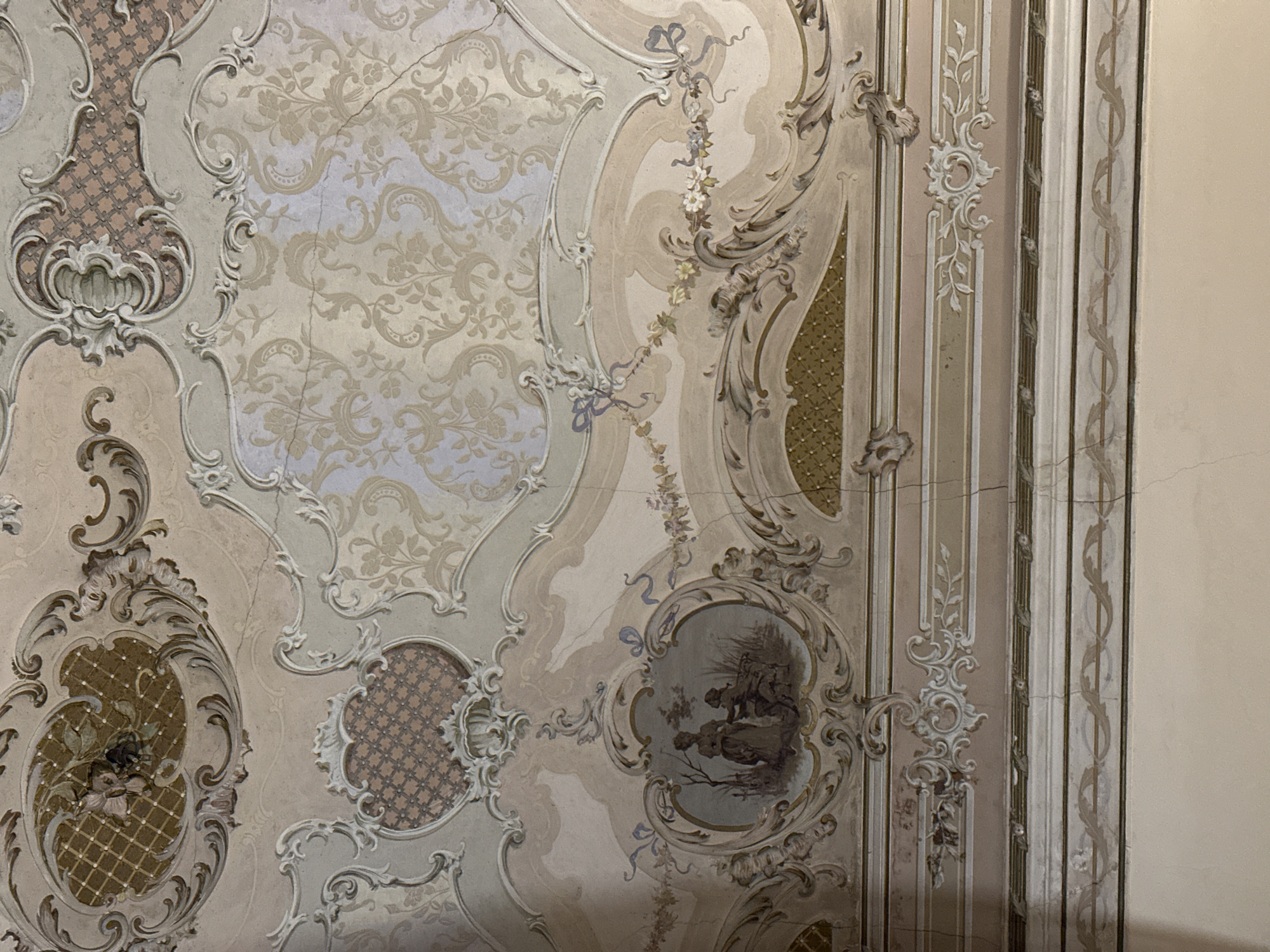 The image shows an ornate ceiling in what appears to be an interior space. The ceiling is decorated with intricate plasterwork or painted designs in muted colors of beige, gray, and lavender. The designs include scrolls, floral motifs, and geometric patterns, suggesting a Baroque or Rococo style. There are also oval medallions with scenes or patterns within them. Cracks and imperfections are visible on the surface, indicating age and possible deterioration.
Based on the presence of this kind of artwork and decoration, the photograph was most likely taken in a historical building, perhaps a church, palace, or museum, in Catania, Italy.