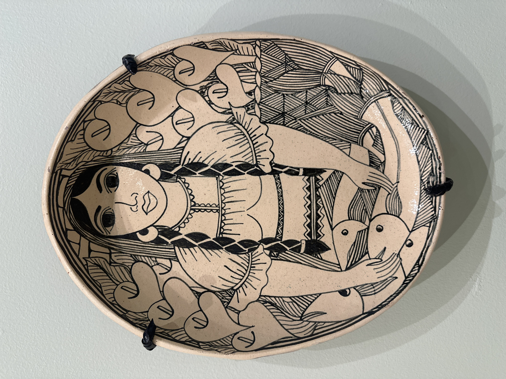 The image shows a round ceramic plate with a cream-colored background and black line drawings. The plate depicts a stylized figure of a woman lying horizontally across the center. She has a large head with prominent eyes, lips, and long hair. Her torso is adorned with geometric patterns, and her arms extend towards either side, surrounded by stylized birds and plants. The rim of the plate is decorated with similar motifs. The plate is attached to a light blue or green wall by two dark-colored fasteners or hangers, positioned on opposite sides of the plate's edge. The overall style of the artwork suggests a folk art or artisan craft tradition, likely from Mexico, although the location in Mexico City specifically cannot be definitively stated.