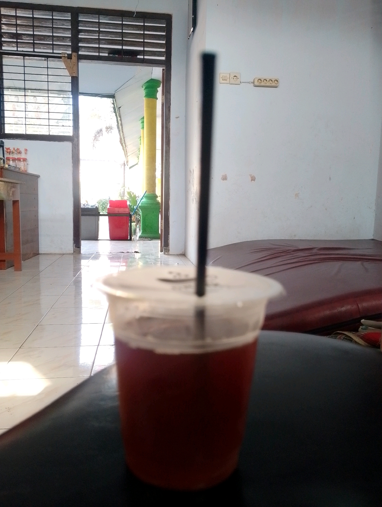 The image depicts an interior scene in what appears to be a modest dwelling or common area in Papungan, Indonesia. In the foreground, a plastic cup filled with a dark red beverage, likely iced tea, is placed on a dark surface, possibly a couch or chair. A black straw extends from the cup. The background reveals a tiled floor reflecting sunlight streaming through an open doorway. Beyond the doorway, a red trash bin and a gray bin are visible, along with two colorful pillars, one yellow and one green, suggesting an outdoor or semi-outdoor space. The wall to the right of the frame features electrical outlets and light switches. A large, dark red mattress or padded surface is situated against this wall. The overall impression is one of a casual, everyday setting.