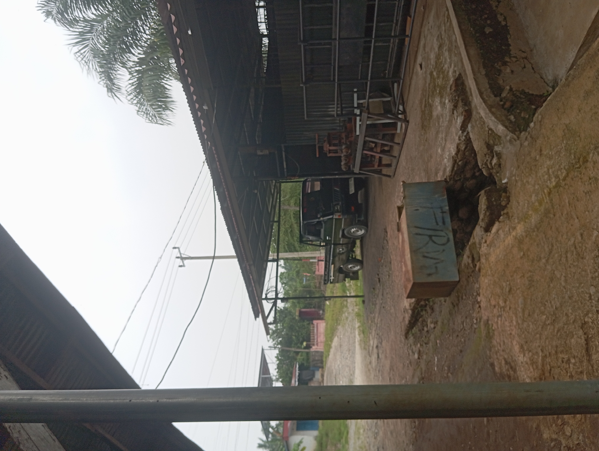 The image depicts an outdoor scene in Desa Retak Mudik, Indonesia. In the foreground, a large grey pole stands to the left, obscuring part of the view. Beyond it, a dirt and gravel path leads into the mid-ground. To the right of the path, a dark-colored utility truck is parked under a covered structure with a corrugated metal roof supported by metal posts. The truck is facing towards the right. In front of the truck, there are some metal racks and machinery. Further back, to the left of the path, is a small building with a white facade and a blue door, and another structure with a pinkish fence in front of it. Lush green trees and vegetation are visible in the background. The sky is overcast, suggesting cloudy weather. A concrete structure with a blue rectangular object on top, possibly a culvert or container, is located in the immediate foreground on the right. The overall impression is one of an industrial or storage area within a semi-rural environment.