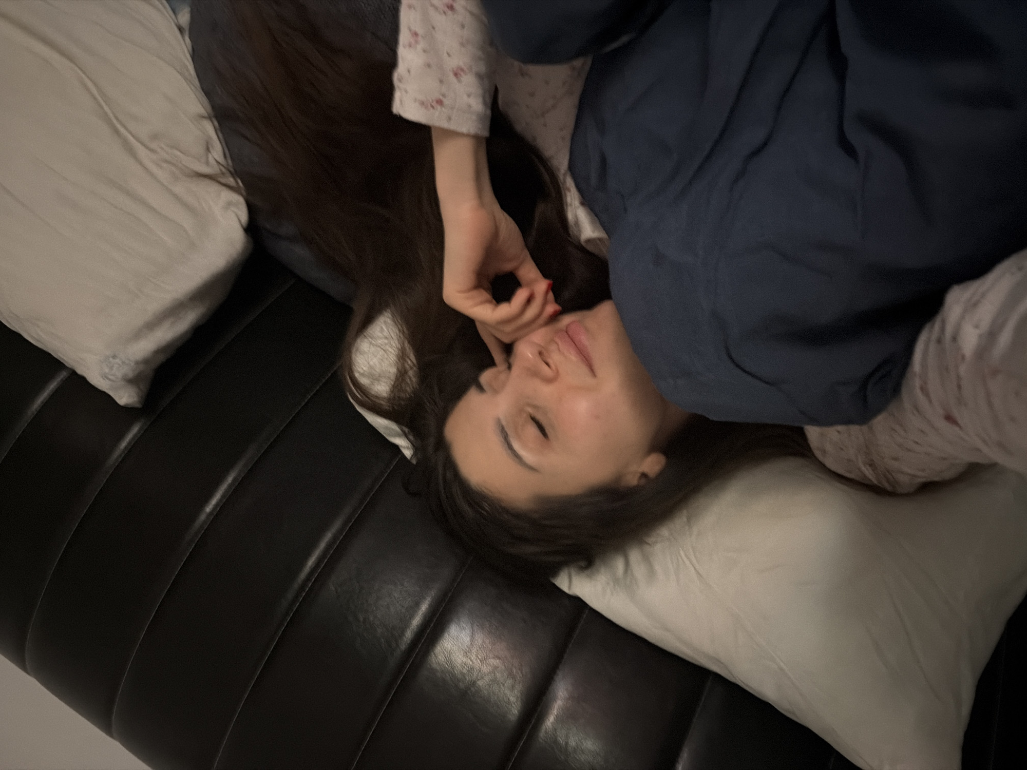 A top-down, close-up image captures a person lying relaxed in an indoor setting. The subject, a female with long, dark hair, is positioned diagonally, resting her head on a white pillow. Her eyes are closed or nearly closed, and a soft, contented smile is visible, suggesting peace, sleep, or gentle awakening.
She wears a white, patterned long-sleeved top, partially covered by a dark blue blanket or duvet. One hand rests near her face, revealing red nail polish on a finger. The background features a black, segmented surface, likely a couch or bed, filling the lower and left parts of the frame. The lighting is dim, indicating a low-light environment, possibly evening or early morning. No specific location cues are visible within the image to suggest Budva, Montenegro, though this is the provided context. There is no visible text.