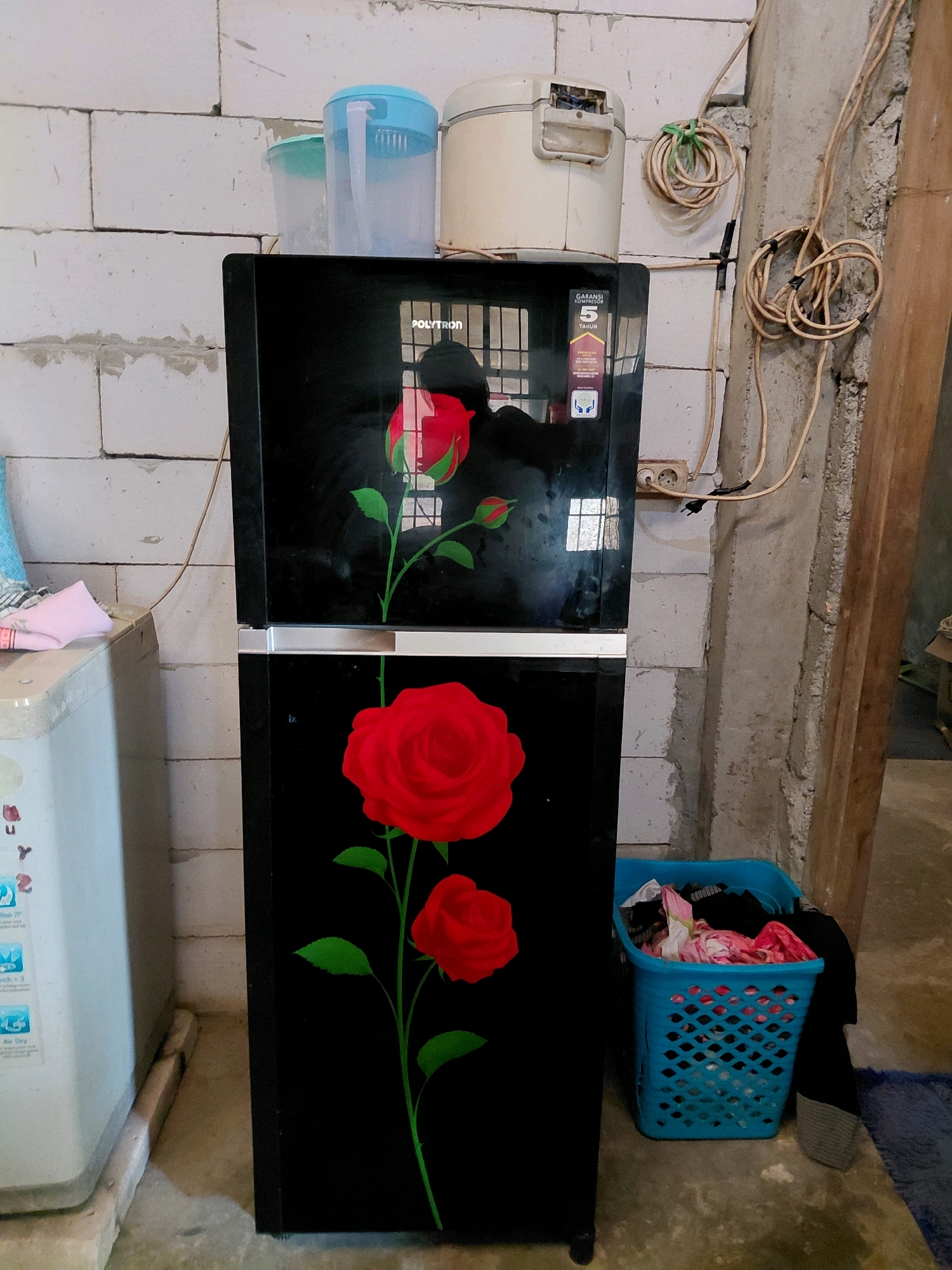 In Dologan, Indonesia, a black refrigerator with a floral design featuring red roses is positioned in a room with unfinished concrete block walls and a concrete floor. To the left of the refrigerator, a light-colored washing machine is visible. On top of the refrigerator, a white rice cooker and two clear plastic water dispensers, one with a blue lid, are present. Electrical cords are routed along the wall behind the refrigerator, connected to a wall outlet. In the foreground to the right, a blue laundry basket is filled with clothes. The scene appears to be indoors, possibly in a residential or utility area.