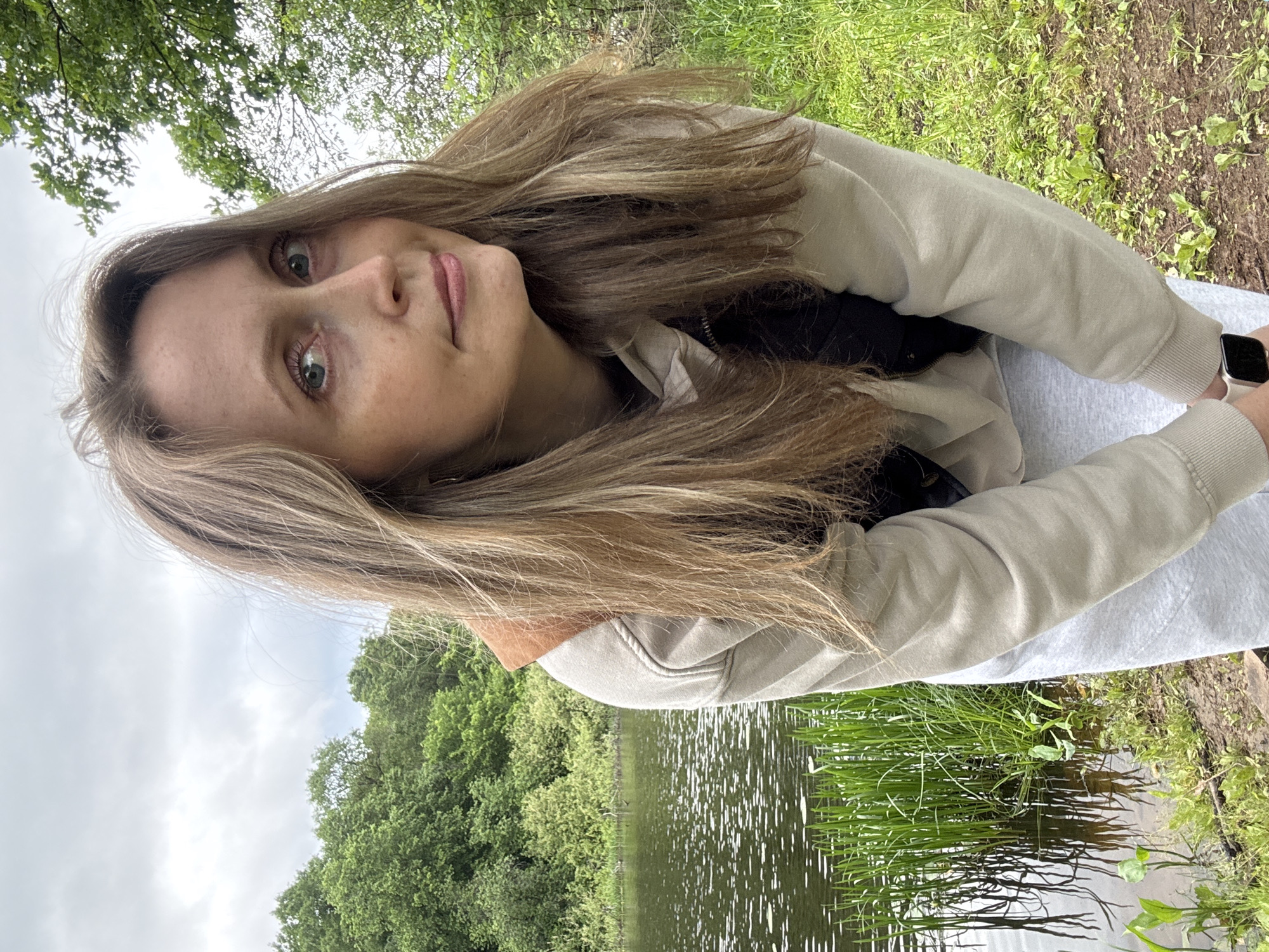 The scene depicts an outdoor setting in the city of Noviy, Russia. A woman, appearing to be in her 30s or 40s, is positioned in the foreground, leaning forward with her torso bent towards the viewer. She looks directly into the camera with a neutral to slightly contemplative expression; her eyes are light blue-green. Her long, light brown hair, with blonde highlights, is parted on the side and falls over her shoulders and chest, appearing slightly windblown. She wears a beige hooded sweatshirt over a dark, possibly black, vest or jacket, and light grey sweatpants. A light-colored smartwatch with a dark screen is visible on her left wrist. Behind her, to the left, is a body of water, likely a lake or pond, with ripples on its surface. Green reeds and taller aquatic plants grow along the water's edge, reflecting in the calm water. Beyond the water and extending to the right, a dense line of various green deciduous trees forms the background. The sky above is overcast with a uniform layer of grey clouds. The woman stands on an uneven dirt bank or path, with sparse green grass and small plants growing in the soil.