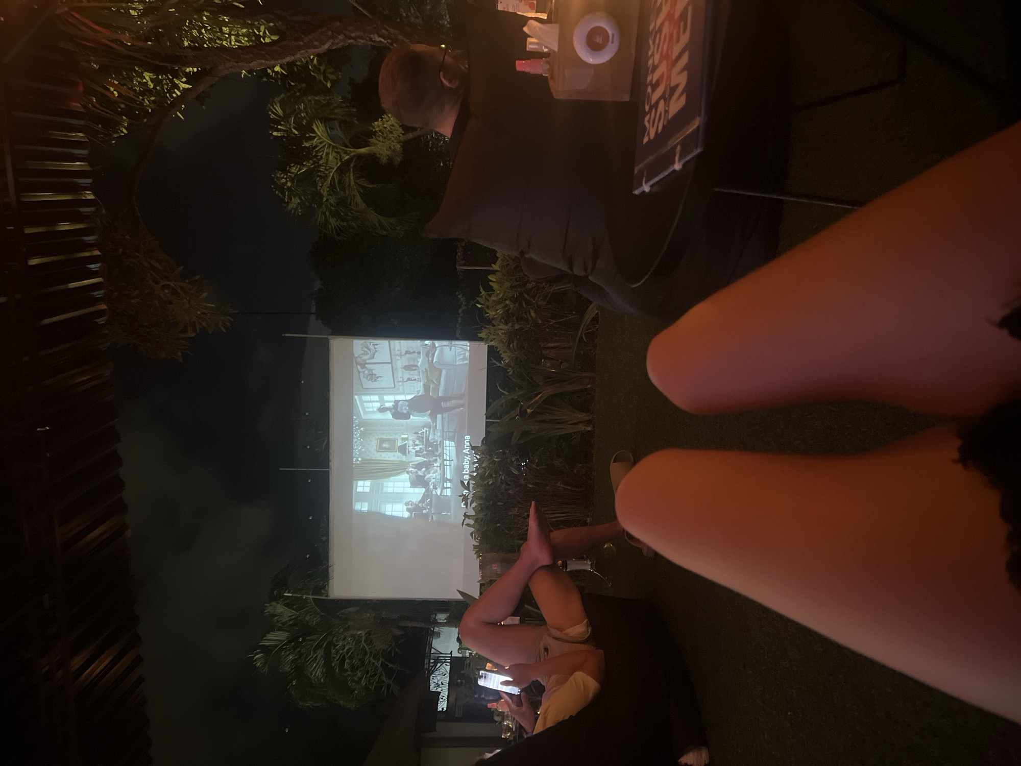 At night in Kuta Selatan, Indonesia, an outdoor movie is being projected onto a screen. A man sits to the side, another person is lying down looking at their phone, and a partial view of two legs is in the foreground. The environment appears to be a garden or patio setting with plants and trees visible.