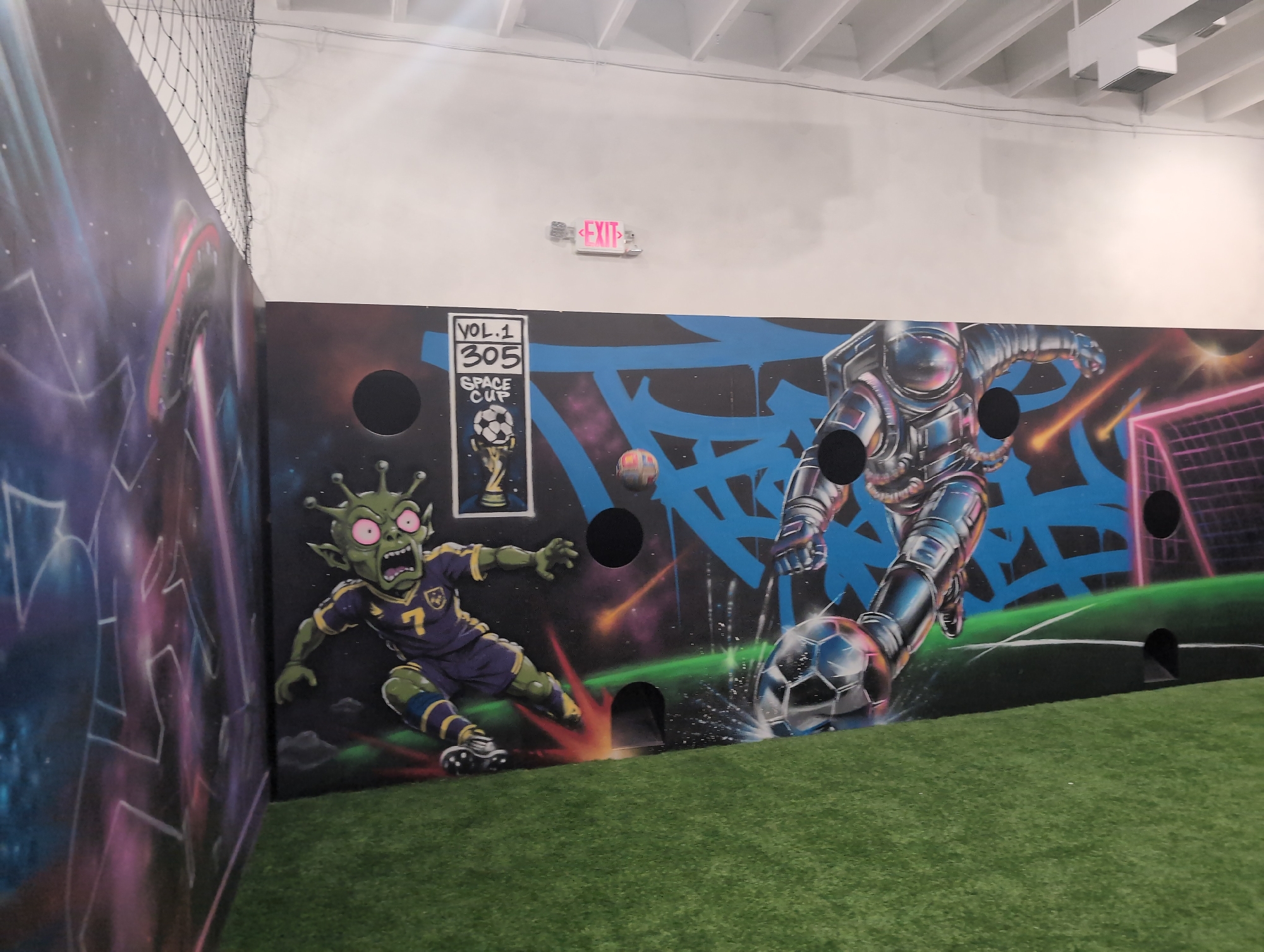 The image shows a mural on a wall in an indoor sports facility. The mural depicts a soccer theme with a cartoon alien wearing a soccer uniform and a robot in a spacesuit kicking a soccer ball. There are also holes in the mural, suggesting it is part of a game. The facility has a green artificial turf floor. An "EXIT" sign is visible on the wall above the mural. The image does not provide enough information to determine the specific location in Miami, United States of America.
