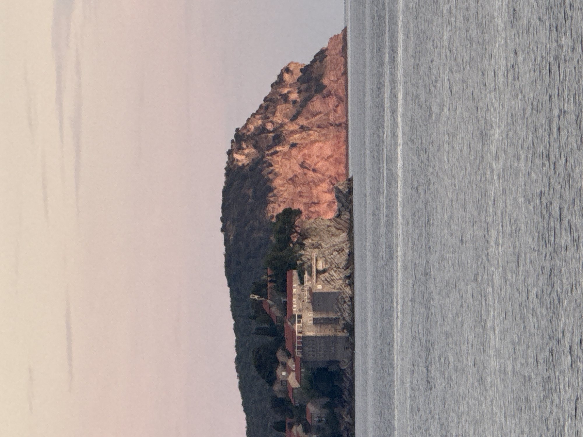 This image depicts a picturesque coastal scene, likely in Rafailovići, Montenegro, captured at what appears to be either sunrise or sunset due to the soft, warm light illuminating the rocky hillside. The orientation of the image is rotated 90 degrees counter-clockwise.
The primary subjects are a rugged, reddish-brown cliff face that dominates the left side of the frame and a calm, grey body of water that fills the right side. Clinging to the lower slopes of the cliff is a collection of buildings with red-tiled roofs, suggesting a small village or settlement. Among these structures, a larger, more imposing building, possibly a fort or an old hotel, stands out with its stone facade and multiple windows. Dark green foliage is interspersed with the buildings and on the steeper parts of the cliff.
The scene is serene, with no visible activity or people. The water's surface is smooth with subtle vertical lines, indicating a tranquil atmosphere. The sky is a pale, hazy pink and white, contributing to the gentle lighting. There are no visible signs of text within the image. The overall impression is one of a quiet, historic coastal settlement nestled against a dramatic natural landscape.