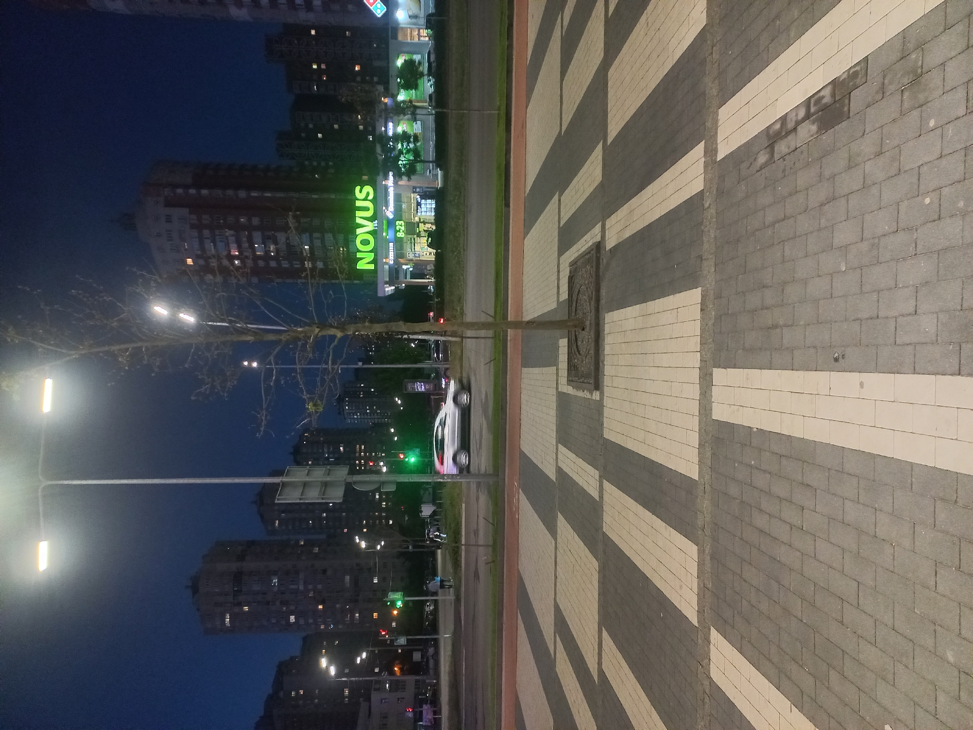 Here's a description of the image:
This is a night scene taken at a street level, most likely in Kyiv, Ukraine. The lighting is primarily from streetlights and building lights, giving the image an overall cool, subdued tone. The dominant element is a wide pedestrian walkway paved with alternating dark and light gray rectangular blocks, creating a striped pattern. A rectangular manhole cover is visible within the walkway.
A tree stands between the walkway and a road. A white car passes by. In the background, several high-rise buildings are visible, their windows lit, indicating residential or commercial occupancy. The "NOVUS" sign in bright green is visible, identifying a supermarket chain often found in Ukrainian cities. A streetlamp shines near the left edge of the frame.