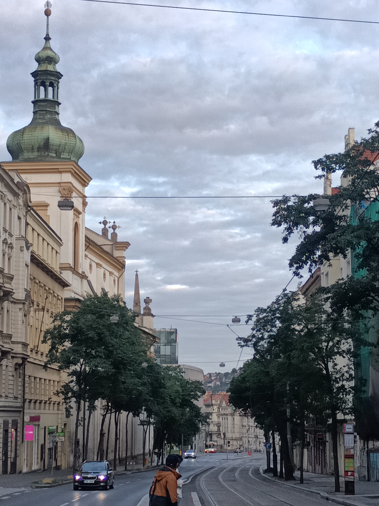 This is a street-level, medium-shot, full-frame photograph taken in Prague, Czechia, likely on a cloudy day. The image captures a view looking down a wide, straight street lined with historic buildings and trees.
On the left, a pale yellow building with intricate architectural details, including arched windows and decorative cornices, dominates the foreground. A church spire with a green copper dome and a pointed finial rises above this building. Further down the street, other ornate buildings with detailed facades are visible.
On the right side of the street, mature trees with dense foliage cast shadows on the pavement. Behind the trees, buildings of varying architectural styles are partially visible, some appearing to be older with decorative elements, while others are more modern. A tram line runs down the center of the street.
In the foreground, a black car with its headlights on is driving down the street. A person wearing an orange jacket and a backpack is standing on the sidewalk, facing the street. In the distance, more cars and buildings can be seen, with hills in the far background. The sky is overcast with grey clouds. Overhead, electrical wires stretch across the frame, indicating the presence of public transportation.