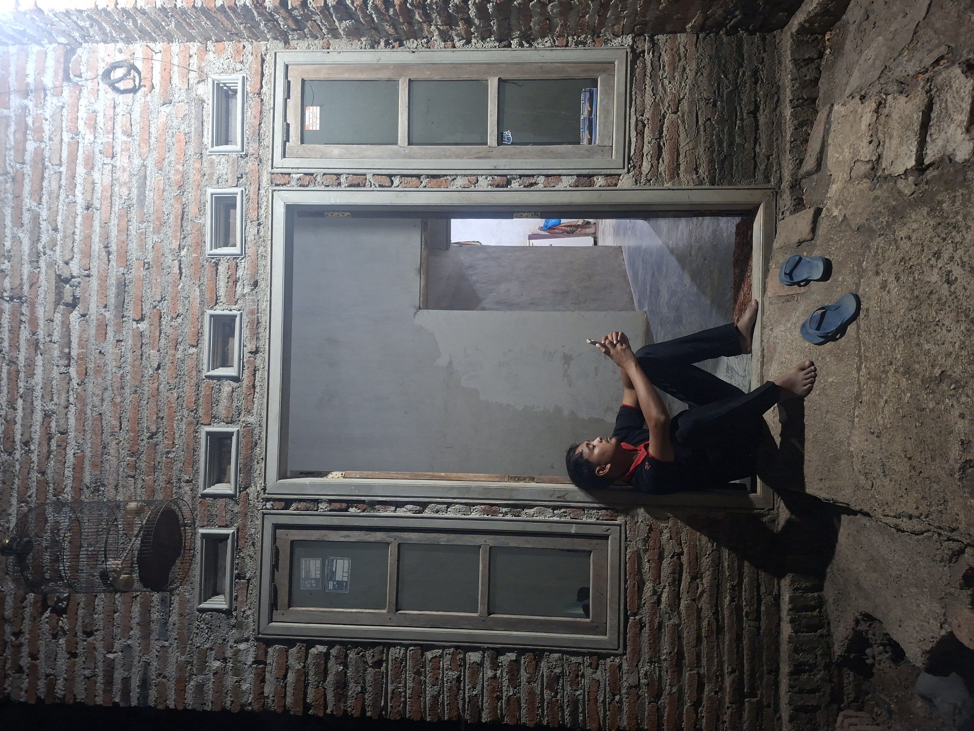 The image shows a man sitting on a concrete step in front of an open doorway. The doorway leads into a dimly lit room. The man is wearing a black polo shirt with a red collar and black pants. He is barefoot and holding a cell phone in his right hand. In front of him, on the step, are two blue flip-flops. The wall behind him is made of rough, red brick. Above the doorway, there are three small, rectangular windows. To the left of the doorway is a wooden-framed window with three panes of glass. To the right of the doorway is another wooden-framed window with three panes of glass. A birdcage hangs from a wire attached to the brick wall above the left window. The scene appears to be an outdoor area of a building in Tongaswetan, Indonesia.