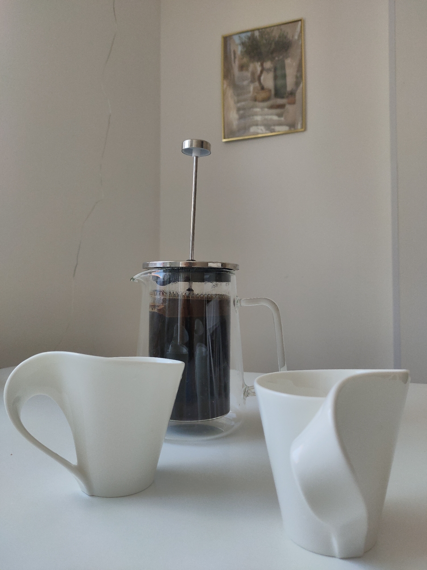 The image depicts a still life composition on a white surface, likely a table. In the foreground, two white ceramic cups with a unique, sculptural design are placed. The cups have a smooth, matte finish and their handles are integrated into their flowing forms. Behind the cups, a glass French press filled with dark liquid, presumably coffee, is centrally positioned. The French press has a silver lid and plunger mechanism. The background consists of a lightly textured, off-white wall. Hanging on the wall, slightly off-center to the right, is a small framed painting with a golden frame. The painting appears to be a landscape with an olive tree, stone steps, and a building. The overall lighting is soft and diffused, suggesting an indoor setting. There is no indication of the specific location within Zrenjanin, Serbia, as the image lacks any identifiable landmarks or contextual clues specific to that city.