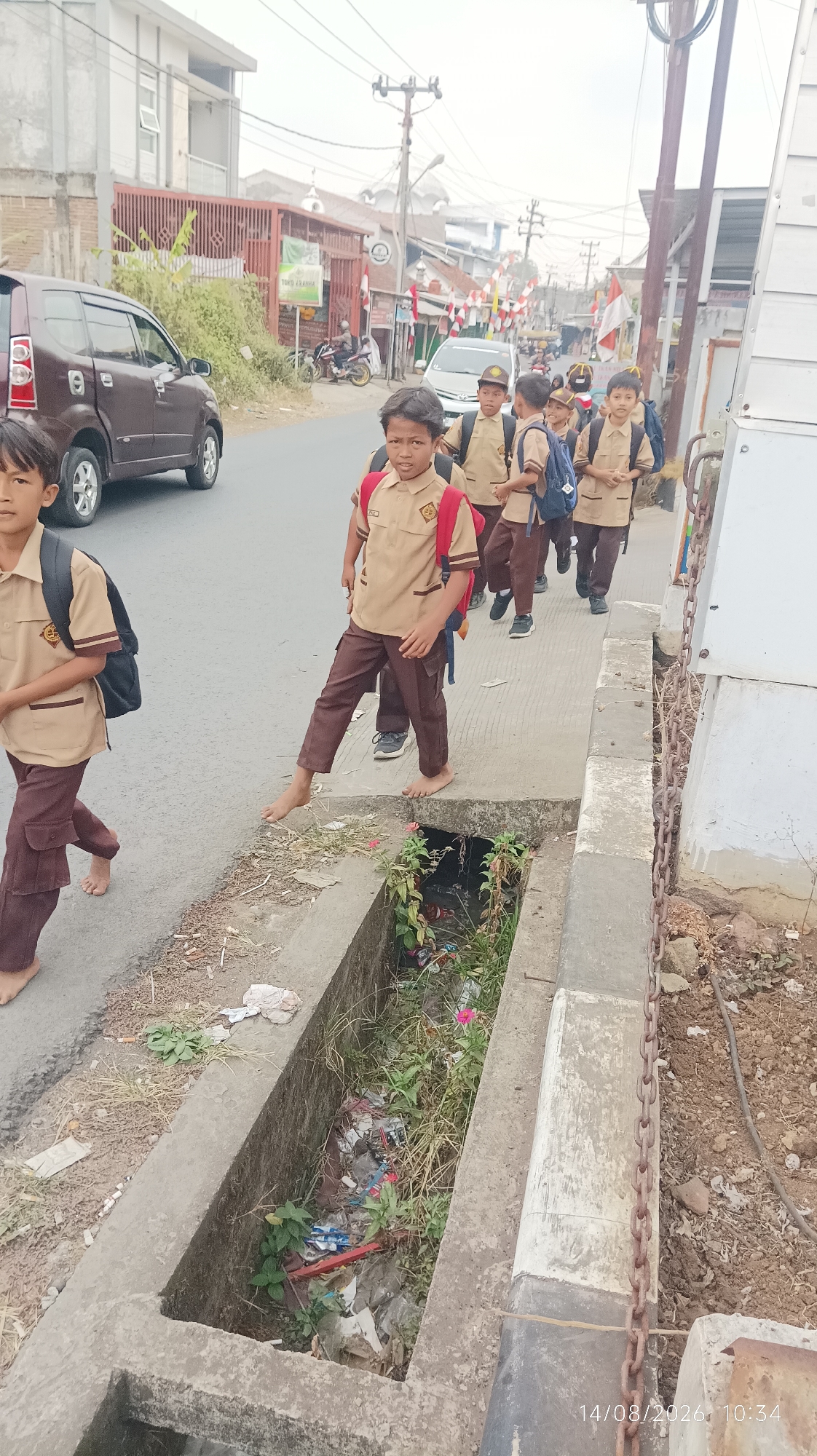 The image depicts a group of young school children in uniform walking along a street in Tasikmalaya, Indonesia. The children are wearing tan shirts and brown pants, and some are carrying backpacks. They are walking on a concrete sidewalk next to a street with a maroon minivan. A drainage ditch filled with debris and vegetation runs alongside the sidewalk. In the background, there are buildings, trees, and a mosque. The date stamp at the bottom right corner indicates August 14, 2026, at 10:34 AM.