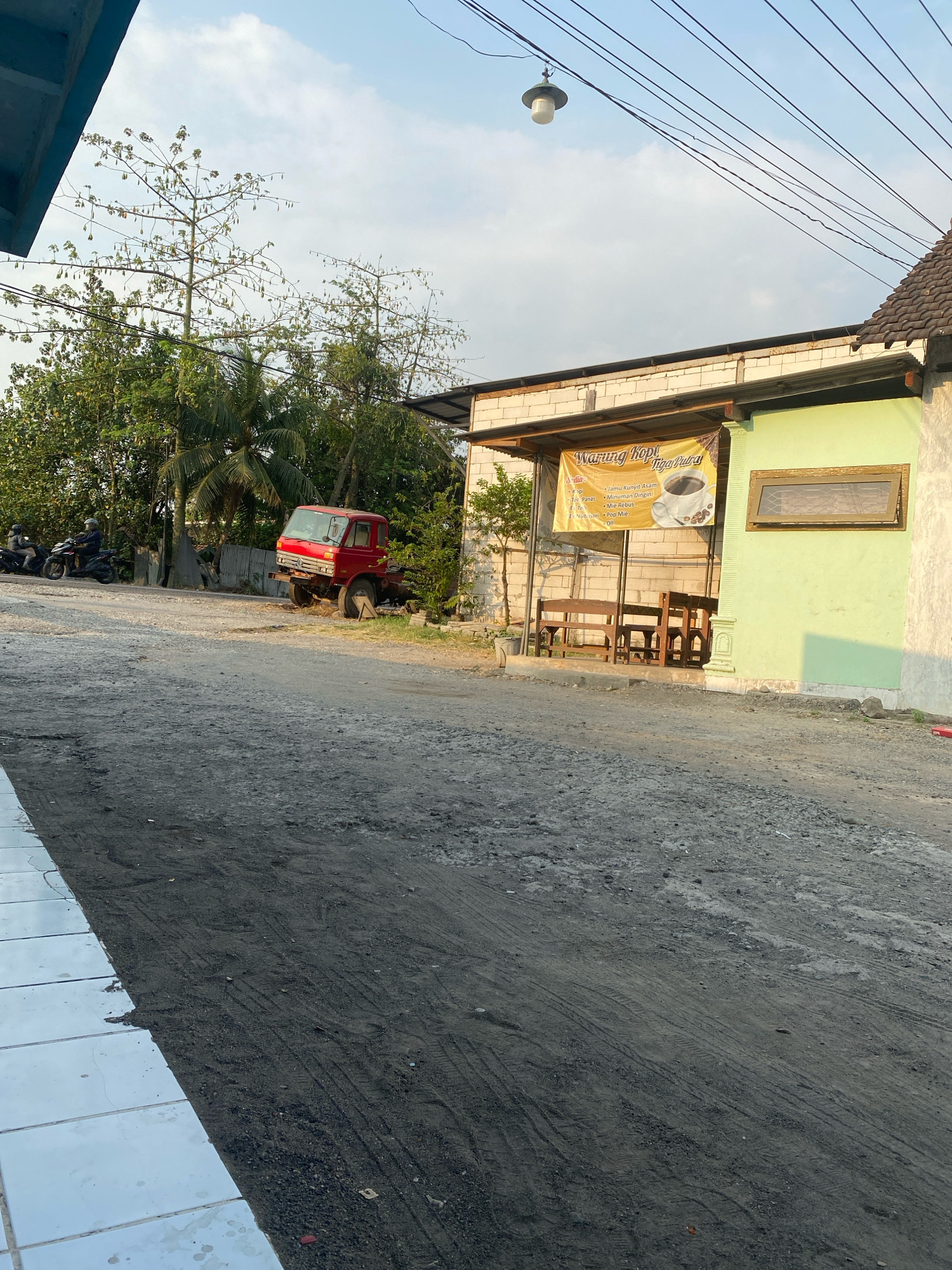 A dusty, unpaved road leads to a small open-air establishment in Kedung Sukodani, Indonesia. The building, constructed with white cinder blocks and topped with a dark tile roof, has a pale green painted section on the right with a wooden-framed window. A sign above the establishment reads "Warung Kopi Tiga Putra," indicating it is a coffee shop. Underneath the sign, wooden benches and tables are arranged, suggesting an outdoor seating area. To the left of the building, a red truck is parked on the side of the road, its front end slightly raised by a wooden support. Several trees, some with sparse foliage and others more densely leaved, surround the area. In the background, power lines crisscross the sky, and a vintage-style street lamp hangs from one of them. Further to the left, two individuals on motorcycles are visible on the road, moving away from the camera. The ground is covered in loose gravel and dirt, with visible tire tracks. The sky is partly cloudy, with the sun casting warm light, suggesting it is late afternoon or early morning.