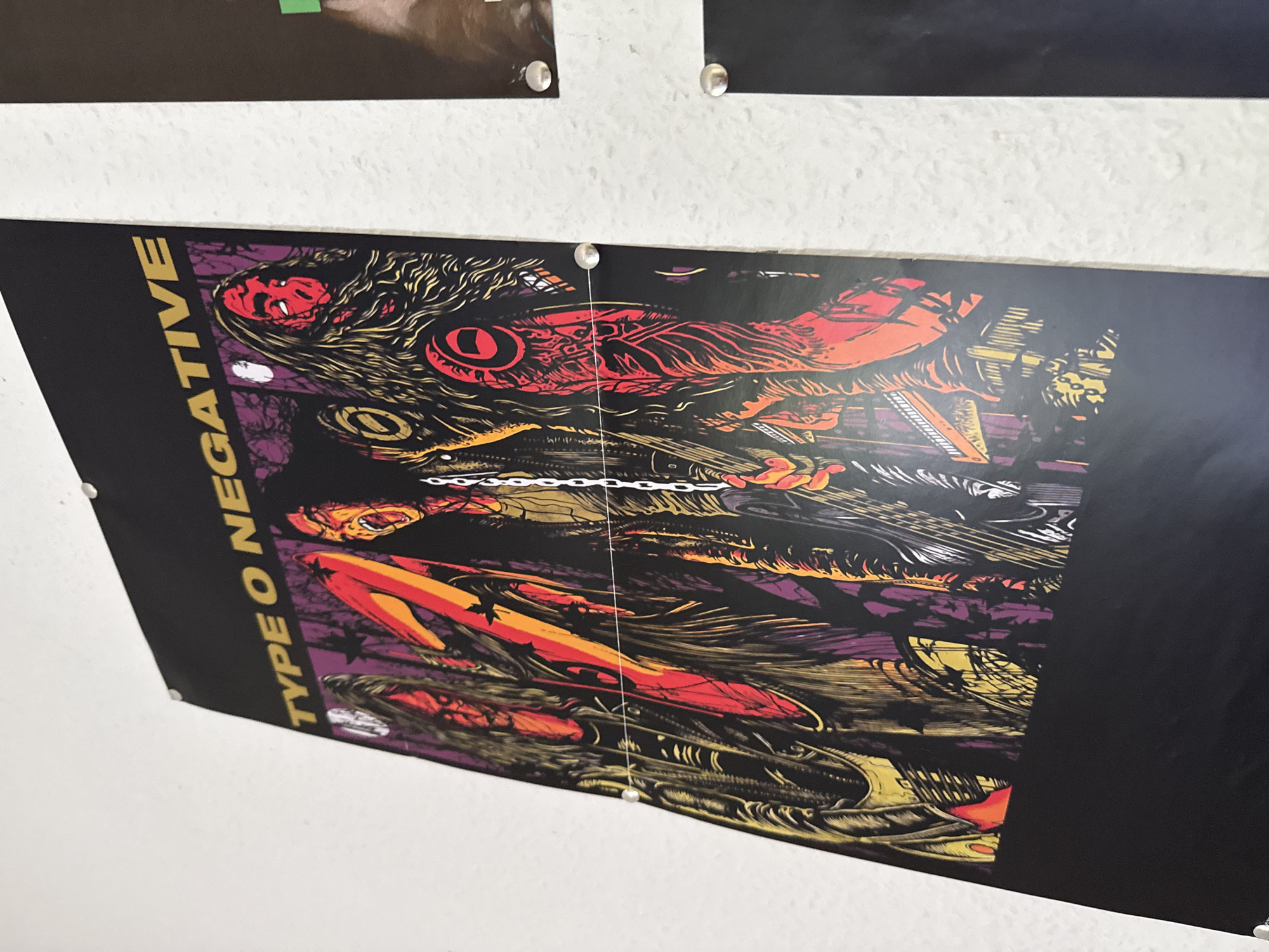 A framed poster of the band Type O Negative is displayed on a white textured wall in an indoor setting, potentially a room or hallway. The poster features a dark, gothic artwork with vibrant red and yellow accents, depicting stylized figures. The poster is attached to the wall with several small silver thumbtacks. The lighting in the image is bright, casting shadows and highlighting the textured surface of the wall and the poster's glossy finish. The image is taken at an angle, showing the poster from the side and slightly from above. The setting appears to be private rather than public. The city of Bad Sachsa, Germany is not visible in the image and cannot be inferred from the context provided.