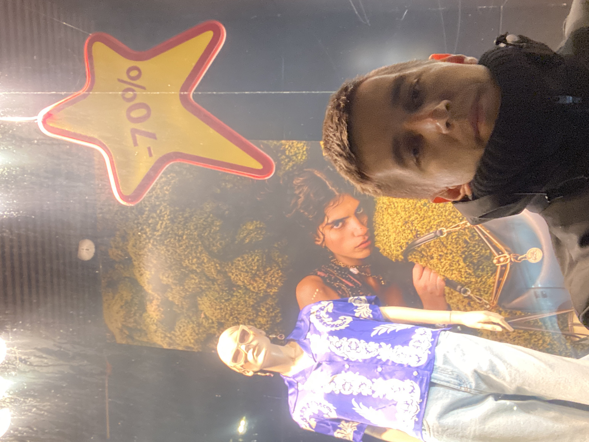 A man is taking a selfie in front of a store window display in Novokuybyshevsk, Russia. The window display features a mannequin wearing a blue patterned shirt and light-colored jeans, and a large advertisement or poster of a woman. A yellow star-shaped sign with "-70%" printed on it is also visible in the window. The man is wearing a black jacket with a high collar. The lighting suggests it is nighttime or a dimly lit interior.