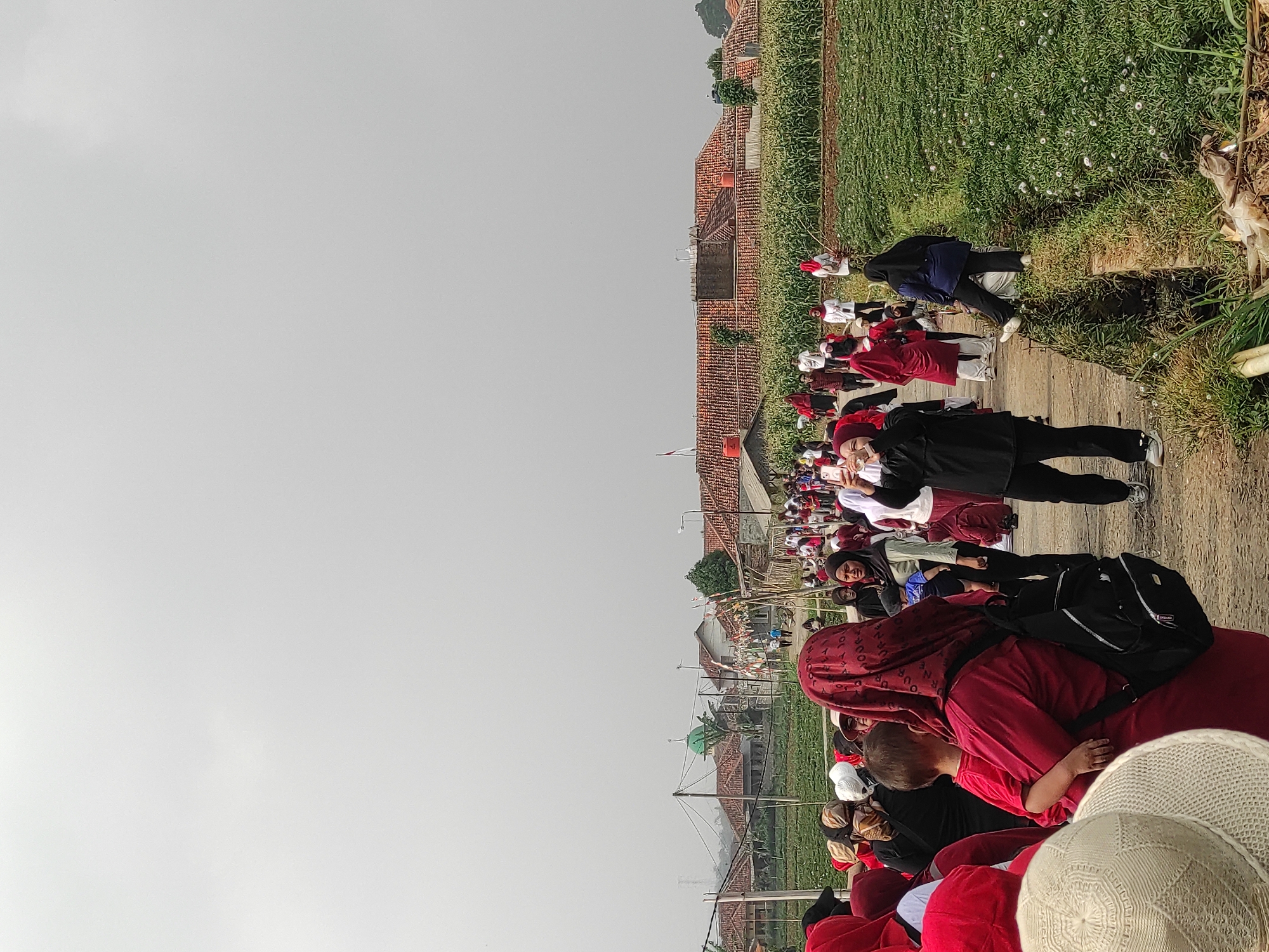 The image depicts a large group of people walking along a dirt path, which appears to be in a rural or semi-rural area. The path is bordered by agricultural fields on the right and houses with terracotta roofs in the background. The sky is overcast.
In the foreground, on the left, a person wearing a wide-brimmed, white hat is partially visible. To their right, a woman in a red hijab and red clothing is holding a young child. She has a black backpack on her back. Behind her, more people in red attire and hijabs are seen.
Further down the path, a woman in a black hijab and black clothing is holding up a smartphone, appearing to take a photo or video. She is wearing black pants and sneakers. To her left, several people in red clothing are walking.
The middle ground shows more people walking, some in red, some in white, and others in darker clothing. They are spread out along the path. In the background, there are several houses with red tile roofs and some vegetation. A green domed building, possibly a mosque, is visible to the left of the houses. Agricultural fields, with tall green plants (possibly corn) and some smaller flowering plants, are prominent on the right side of the path.
The overall scene suggests an event or gathering, possibly a parade or procession, taking place in a village setting. The presence of many people, some in uniform red clothing, indicates a coordinated activity. The rural landscape with houses and fields points to a location outside of a dense urban center. The image does not provide explicit visual cues to pinpoint the exact location within Sumedang, Indonesia, but the architectural style of the houses and the agricultural context are consistent with rural Indonesian settings.