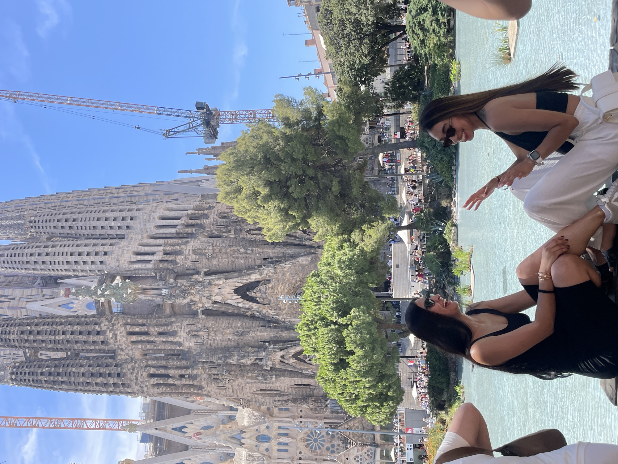 The image captures a sunny day in Barcelona, Spain, with the iconic Sagrada Familia basilica as a prominent backdrop. The basilica, still under construction, is visible in its intricate stone facade, with a large crane positioned nearby, indicating ongoing work.
In the foreground, two women are relaxing by a body of water. The woman on the right is wearing a black tank top and white pants, and sunglasses, smiling and looking to her right. She has a watch on her left wrist and bracelets on her right. The woman on the left, also wearing a black outfit, is seated with her legs crossed, looking up and laughing. Her outfit appears to be a black dress, and she has a black headband or hair accessory.
The scene is lively, with many people visible in the background, suggesting a tourist area or a public gathering. Lush green trees provide a natural element to the urban landscape. The bright sky and clear visibility indicate fair weather. The overall mood is relaxed and joyful, with the subjects appearing to enjoy their time outdoors in a scenic location.