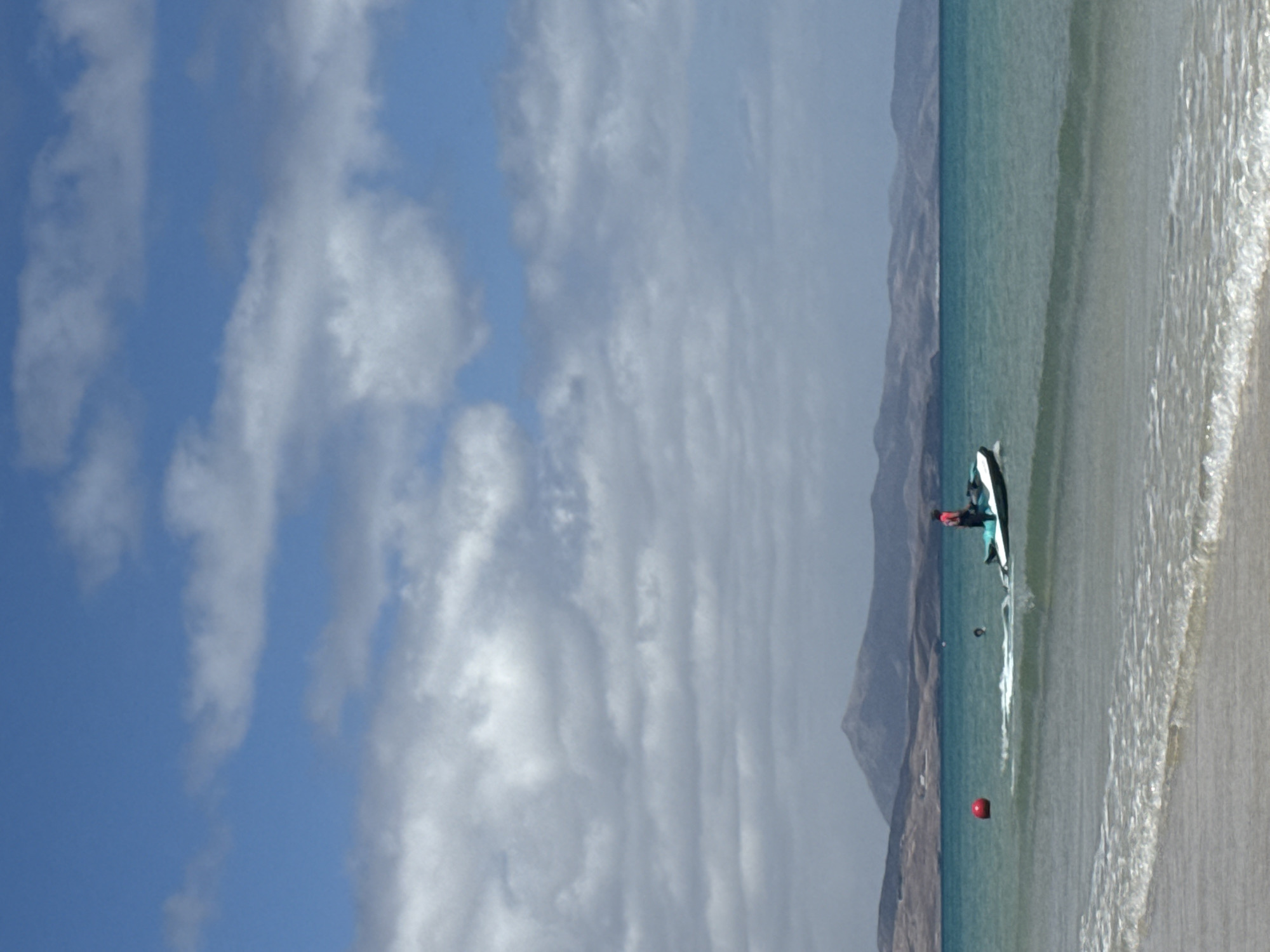 The image shows a beach scene in Pájara, Spain. A person is on a stand-up paddleboard in the turquoise water, near the shoreline. In the water, there is a red buoy floating. Mountains are visible in the background. The sky is blue with many white clouds. The beach has tan sand and small waves are crashing on the shore.