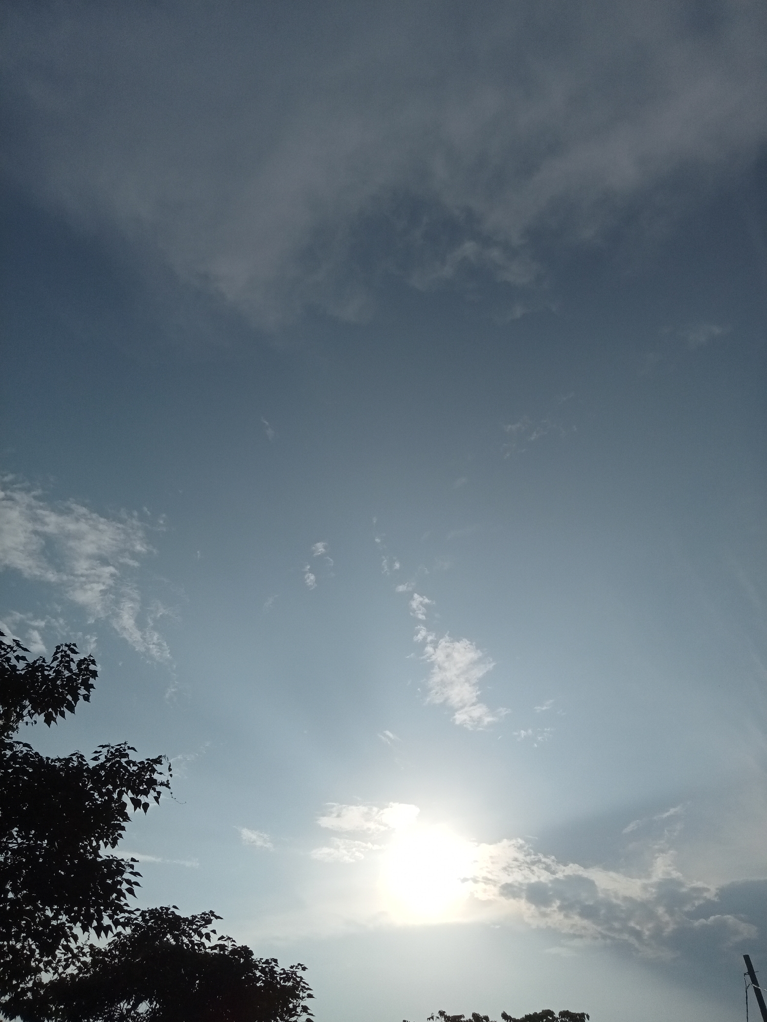 The image depicts a sky with scattered clouds and the sun partially obscured by them. The sun's rays are visible, creating a luminous effect. In the lower left corner, the silhouette of tree branches with leaves stands out against the sky. In the lower right corner, a portion of a utility pole is visible. The overall scene is characteristic of an outdoor environment, likely in a suburban or rural area given the presence of trees and a utility pole, and could be located in Teluk Kenidai, Indonesia. No people or specific actions are discernible in the image.