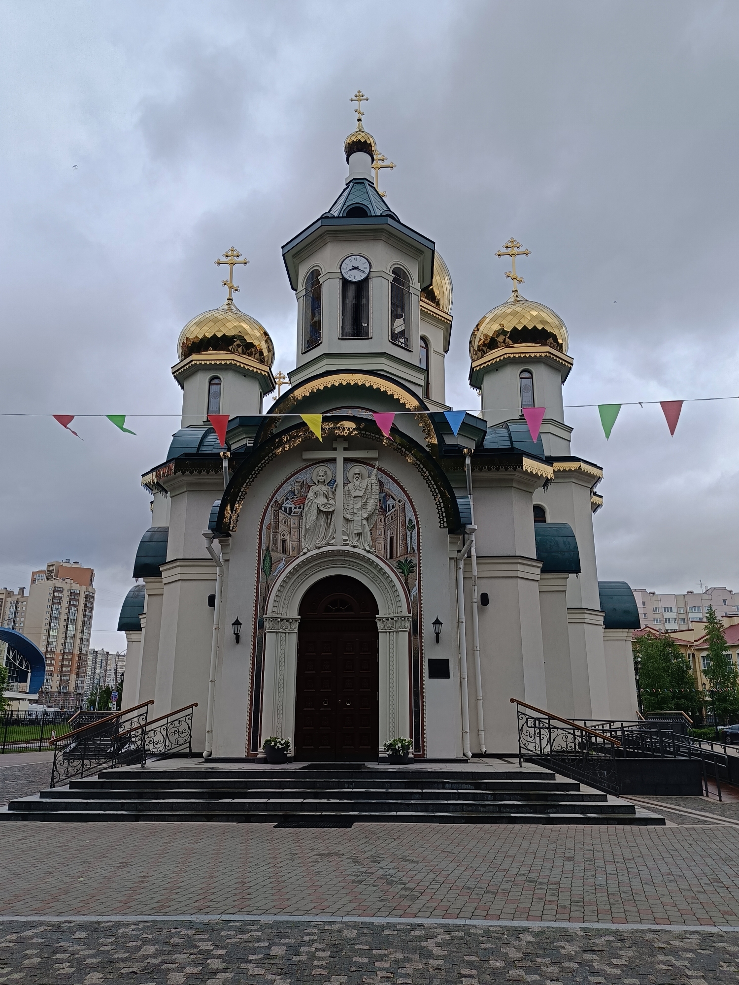 The image depicts a white Orthodox church with golden domes, a brown door, and a clock on its central tower. The church has decorative carvings and paintings above the entrance. Multi-colored flags are strung between the domes. The church is accessed by a set of steps with black railings on either side. The ground in front of the church is paved with multi-colored bricks. In the background are apartment buildings under an overcast sky. The image was taken in Saint Petersburg, Russia.